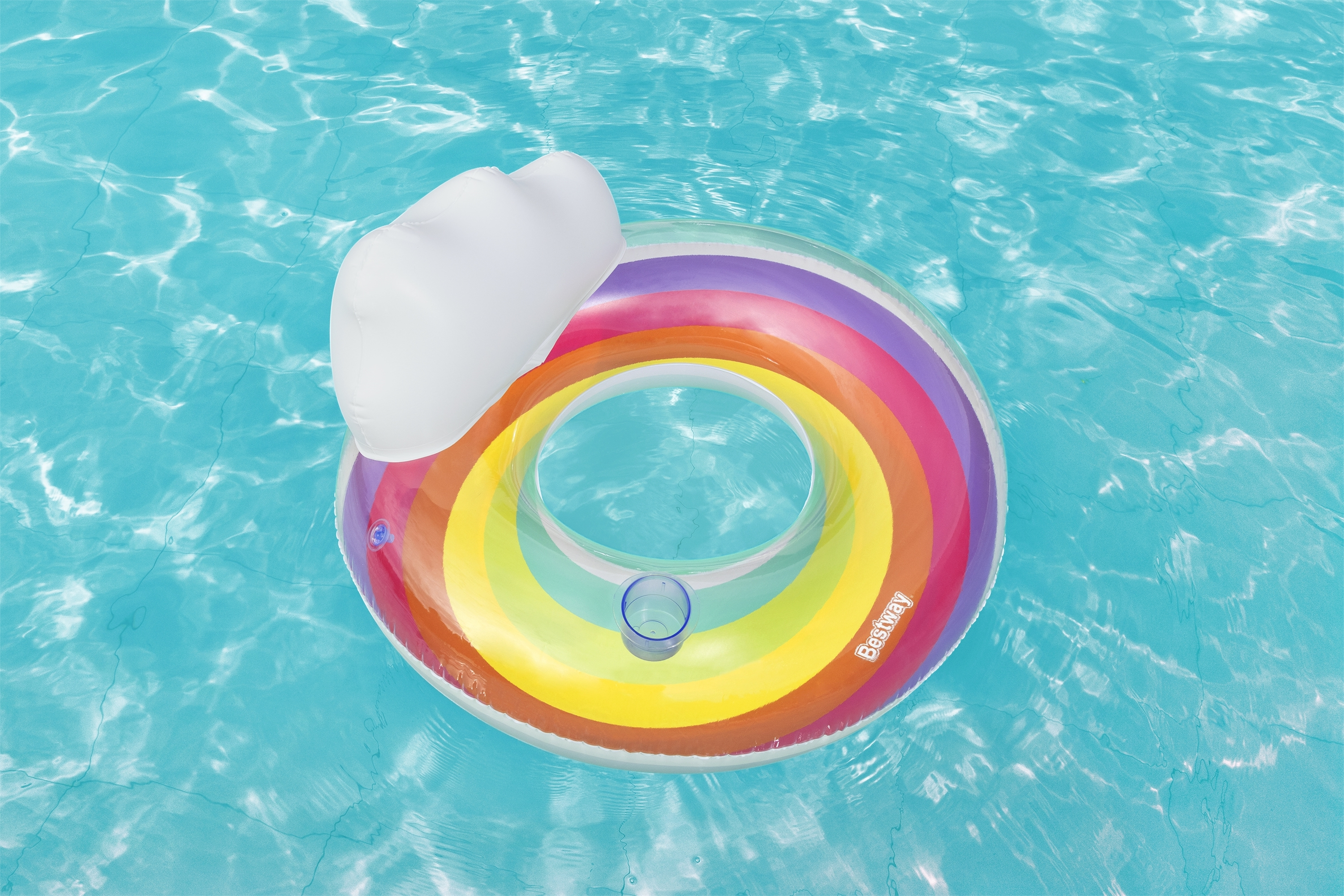 Un flotador inflable de arcoíris con reposacabezas blanco flota en una piscina azul. Un portavasos es visible.