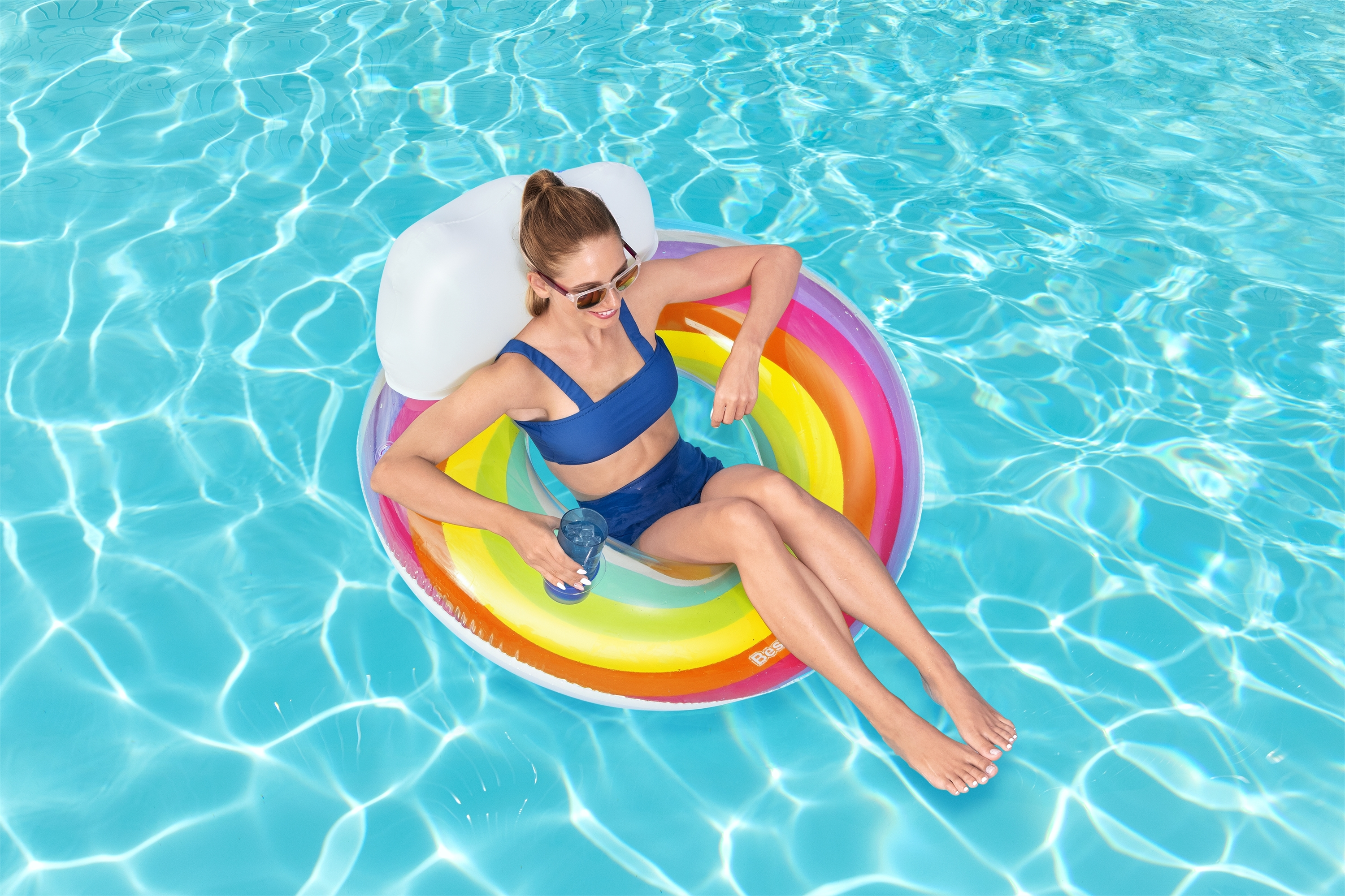 Mujer en traje de baño azul en flotador arcoíris en piscina, con bebida, día soleado.