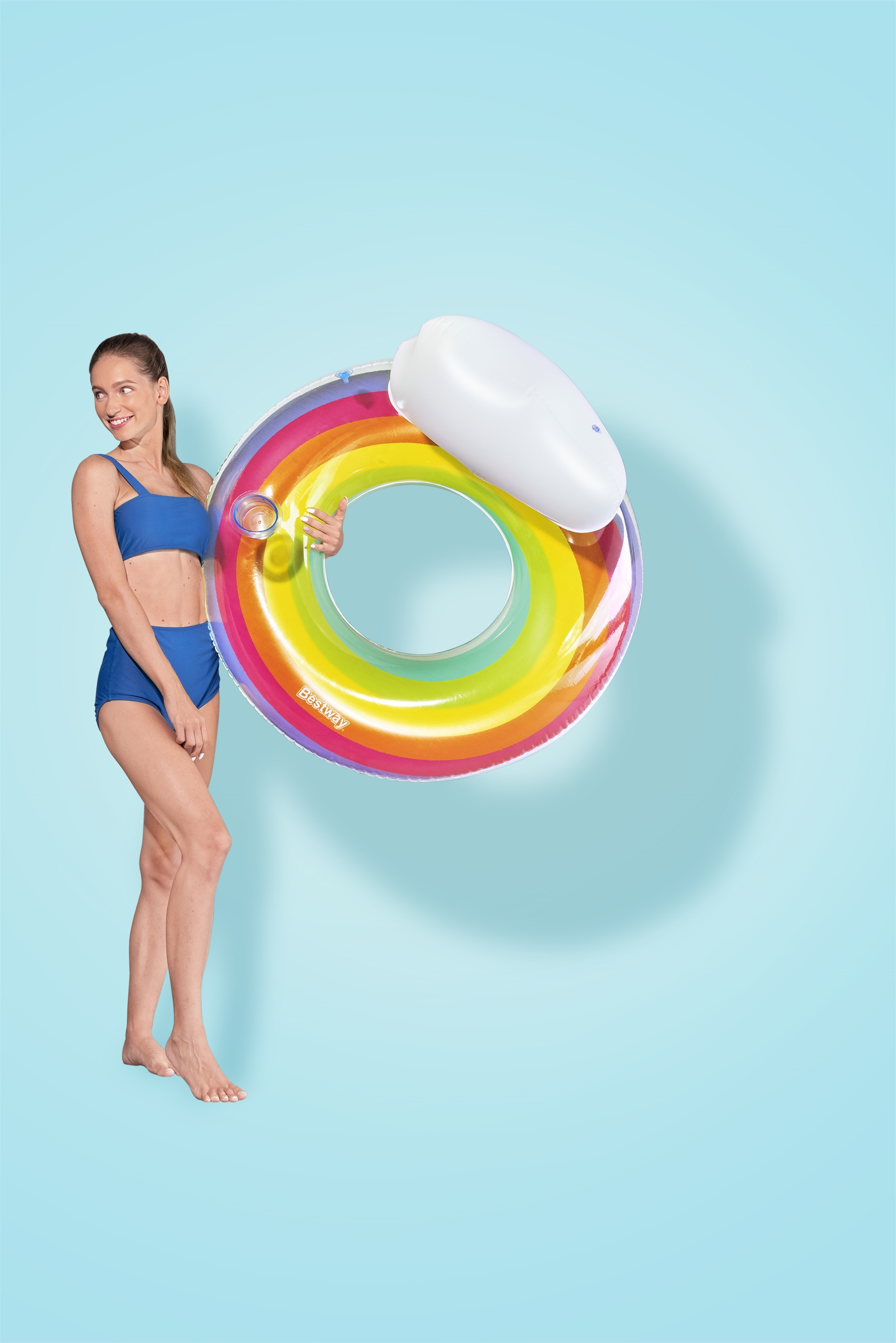 Mujer en bikini azul sosteniendo un flotador arcoíris con almohada blanca sobre fondo azul.