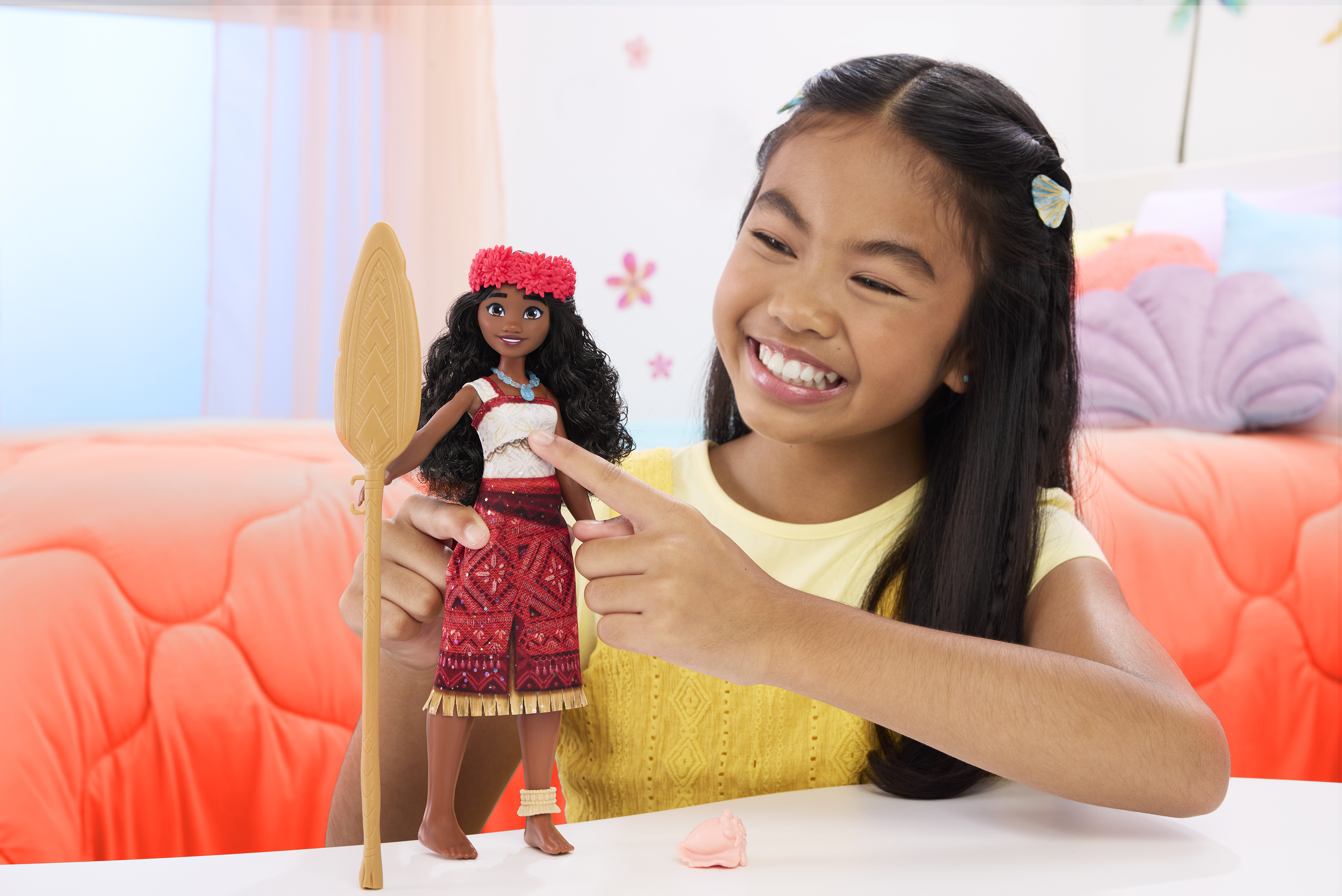 Dziewczyna uśmiecha się, trzymając lalkę. Lalka ma czerwoną sukienkę i trzyma drewniane wiosło. Widoczna jest różowa poduszka.