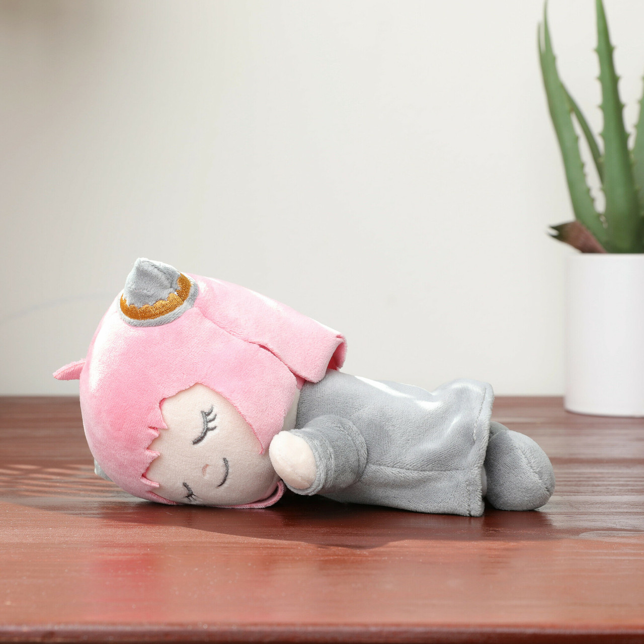 Pluszowa lalka z różowymi włosami i szarymi ubraniami leży na drewnianej powierzchni. Roślina w białej doniczce jest w tle.