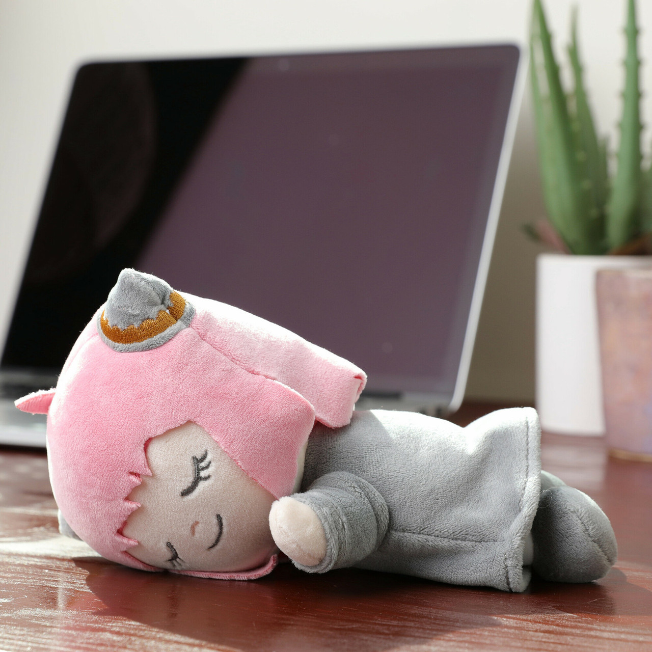 Pluszowa zabawka z różowymi włosami i szarymi ubraniami leży na biurku. Laptop i roślina są w tle.