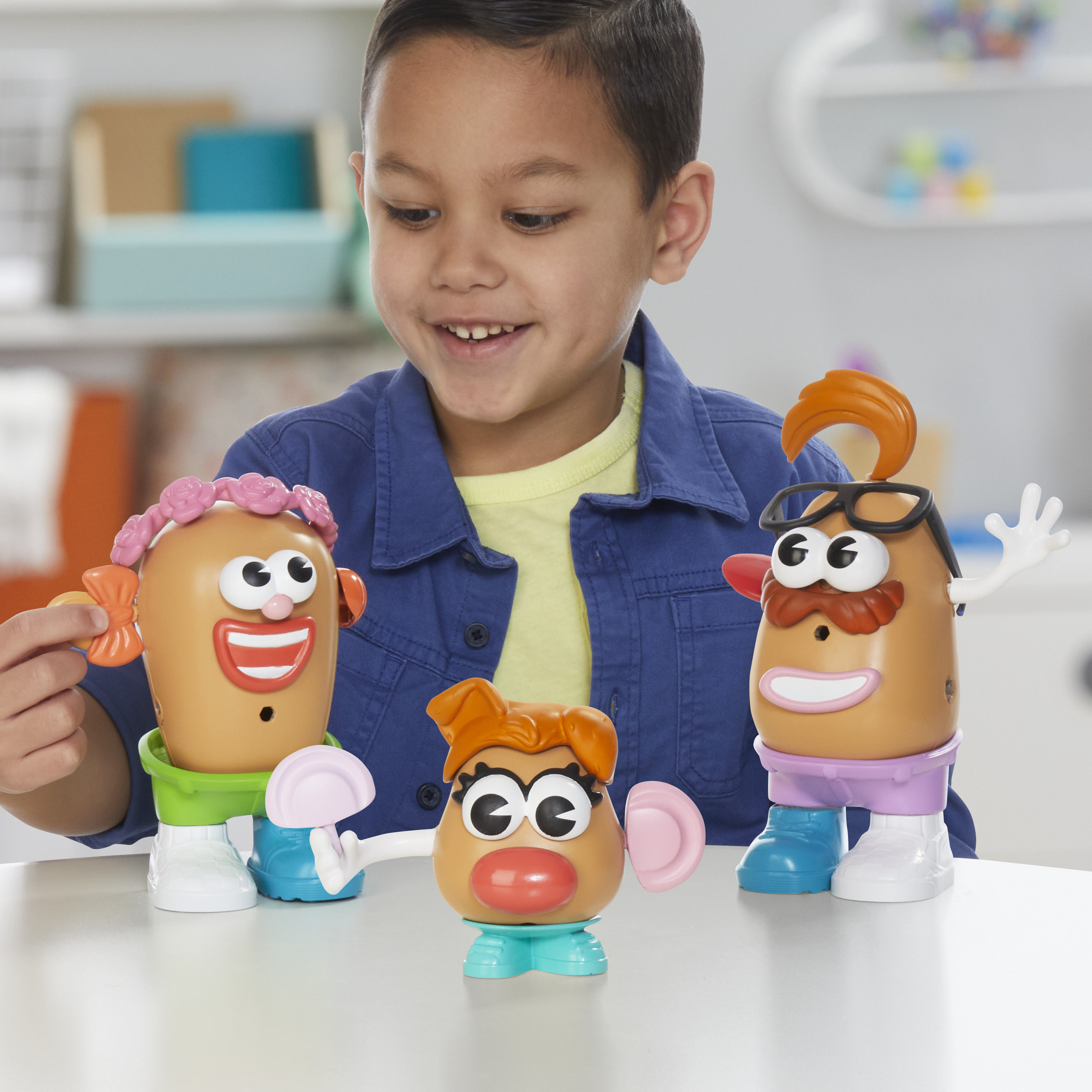 Un niño sonríe a tres juguetes de Mr. Potato Head. Cada uno tiene accesorios diferentes como pelo, gafas y zapatos.