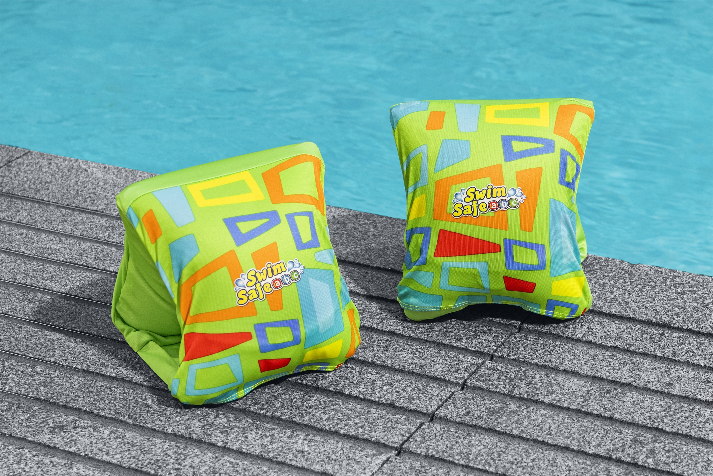 Zwei grüne Schwimmflügel mit bunten geometrischen Mustern auf grauer Steinfläche, am Pool.