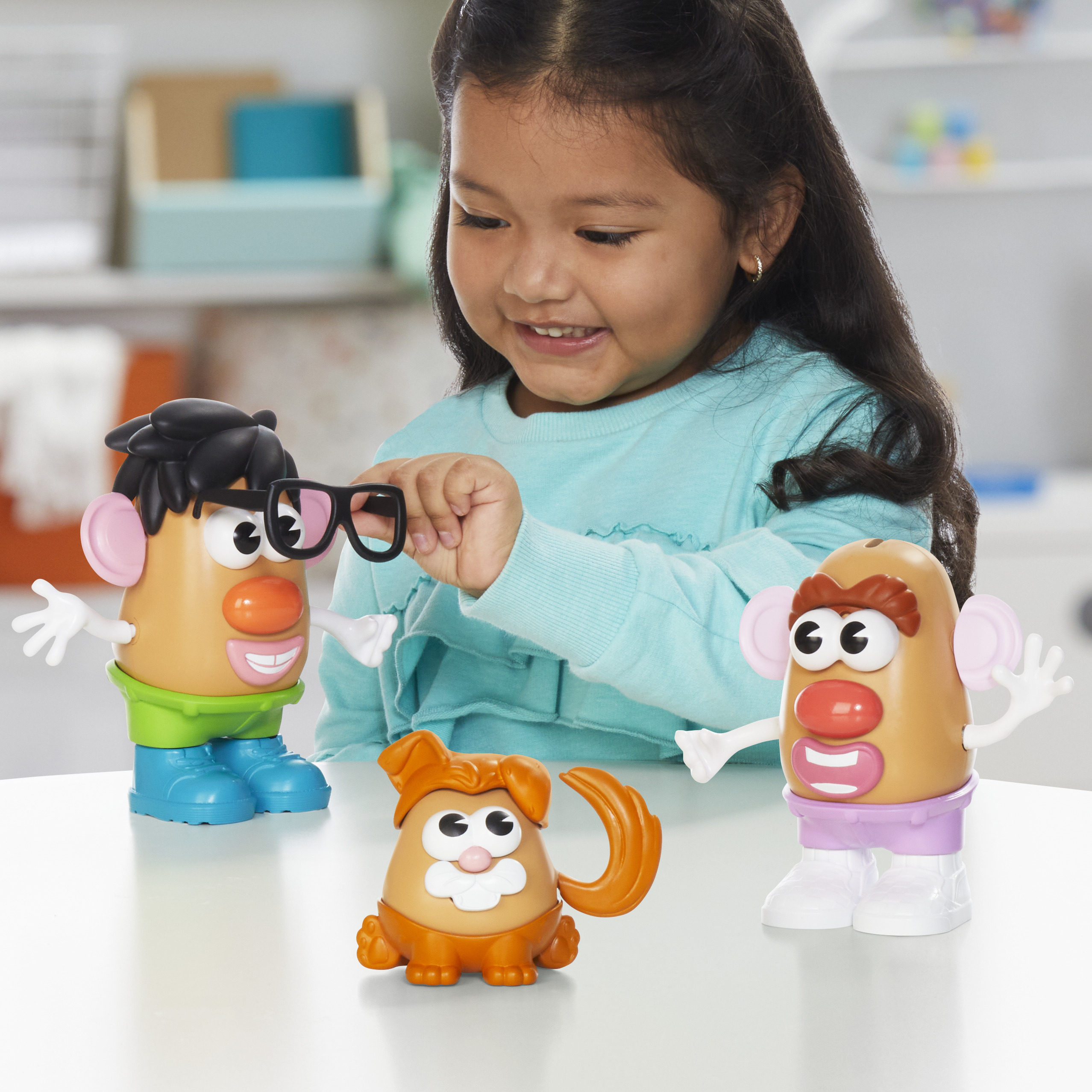 Una niña sonríe a los juguetes de Mr. Potato Head. Uno tiene gafas, otro un perro. Todos están en una mesa blanca.