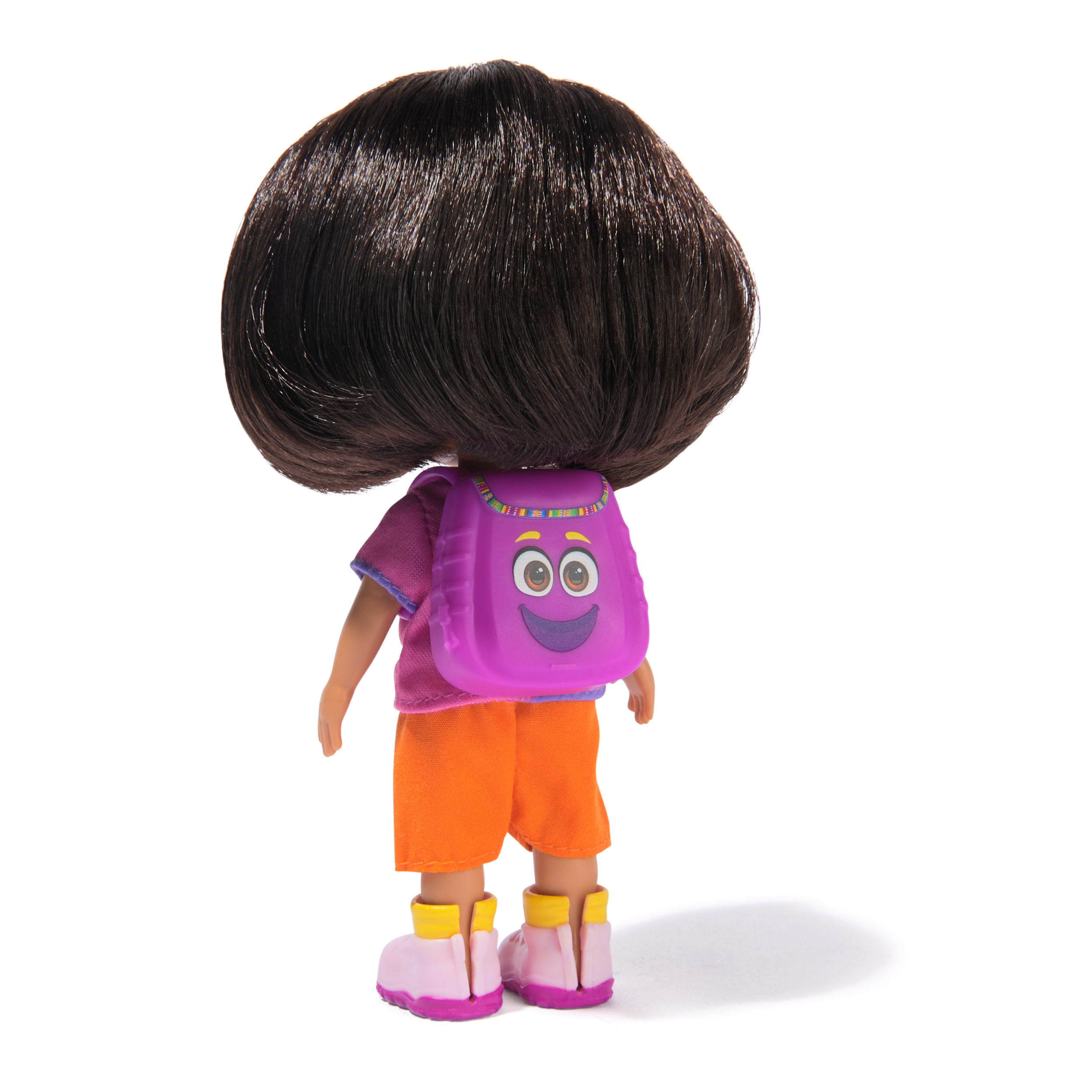 Muñeca, vista trasera, con cabello oscuro, mochila morada con cara, zapatos rosas, pantalones cortos naranjas, camisa morada. Fondo blanco.