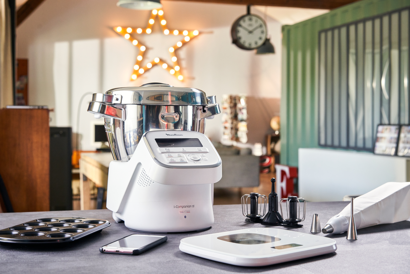 Una escena de cocina con un procesador de alimentos, utensilios para hornear y una luz en forma de estrella. Una balanza y un teléfono están sobre la mesa.