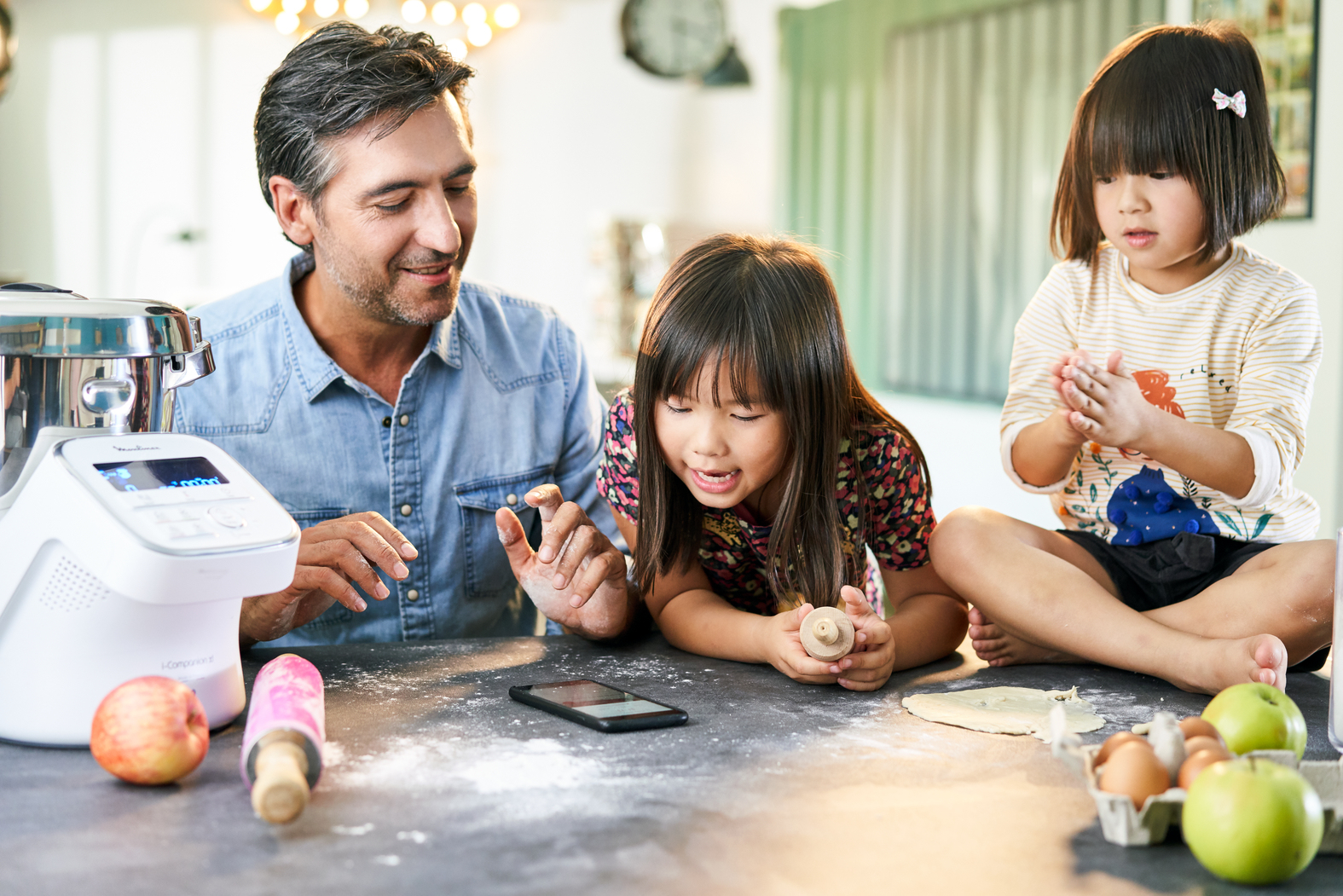 Un hombre y dos niñas hornean. Ingredientes, rodillo y smartphone están sobre la mesa. También hay un procesador de alimentos.