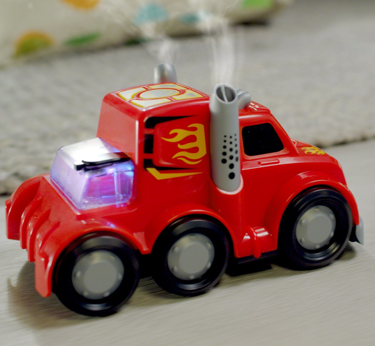 Czerwona zabawka ciężarówka z płomieniami, dymem i czarnymi kołami na szarej podłodze. Ciężarówka ma podświetlane niebieskie okno i szare rury wydechowe.