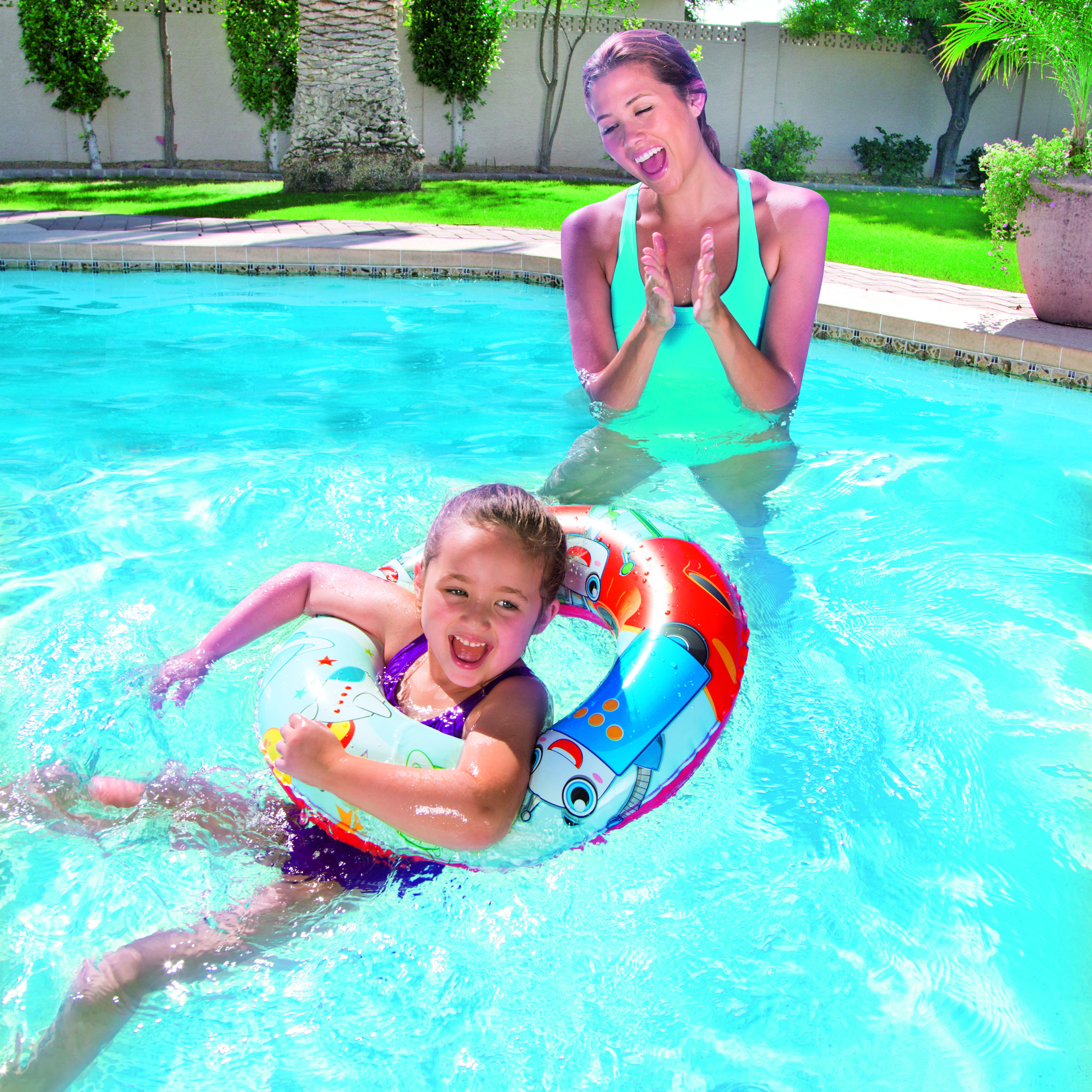 Una niña en una piscina con un flotador sonríe a la cámara. Una mujer anima desde el agua.