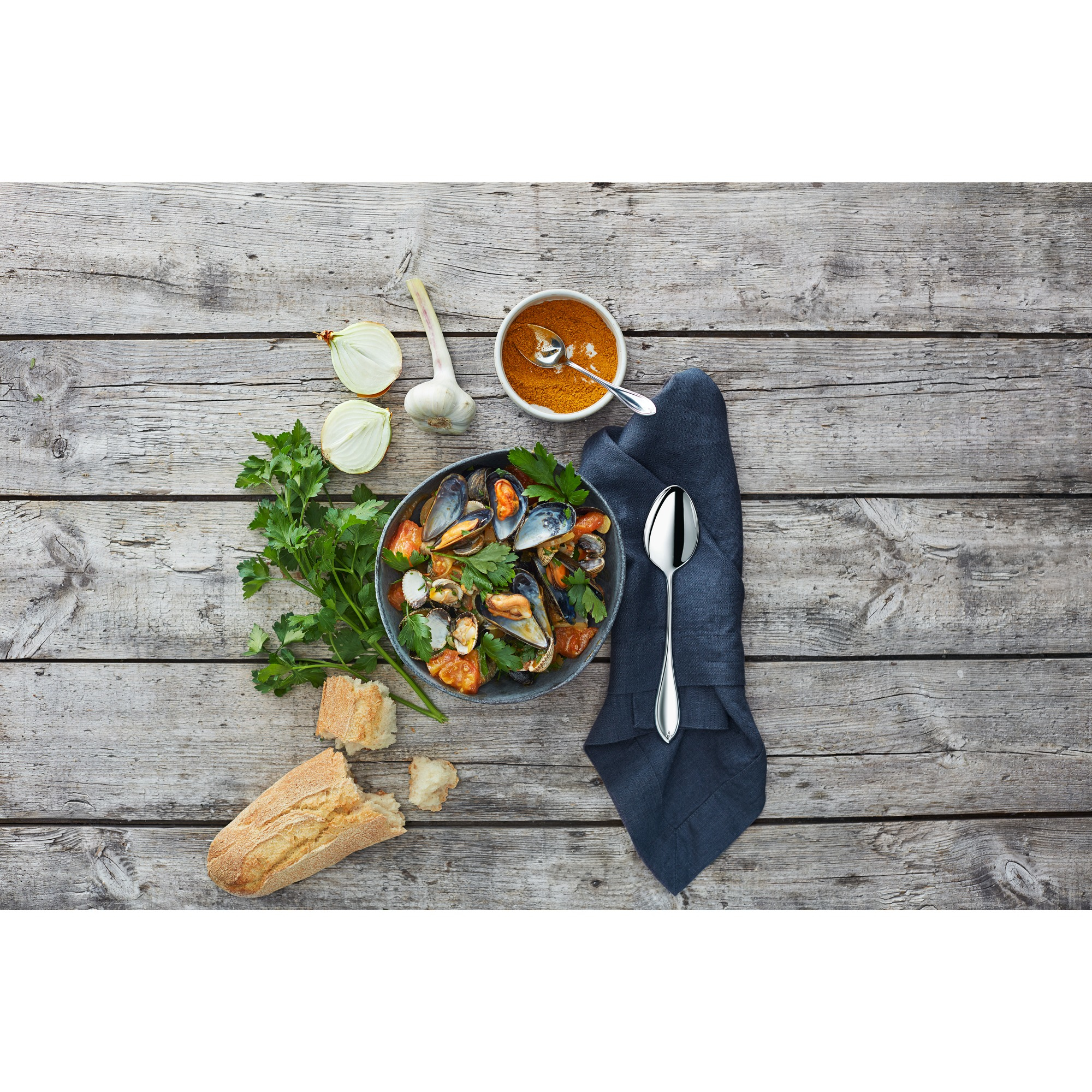 Eine Schüssel Muscheln mit Tomaten und Petersilie, neben Brot, Zwiebel, Knoblauch, Gewürzen, Löffel und Serviette auf Holz.