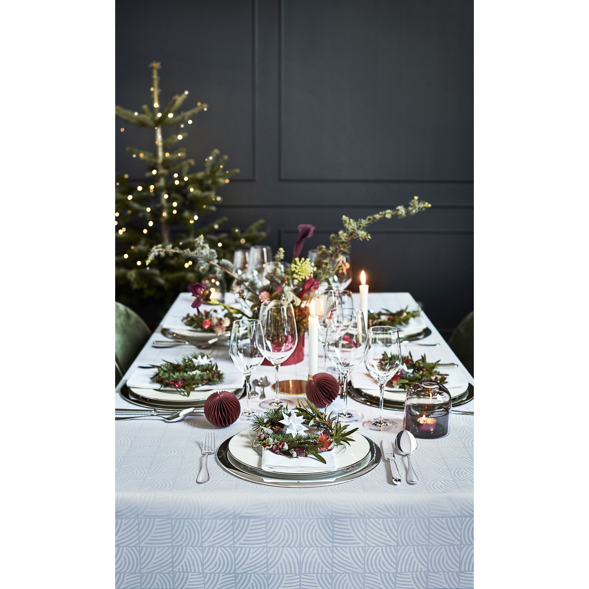 Festliche Tischdekoration mit weißen Tellern, Kerzen, Weingläsern und einem geschmückten Weihnachtsbaum im Hintergrund.