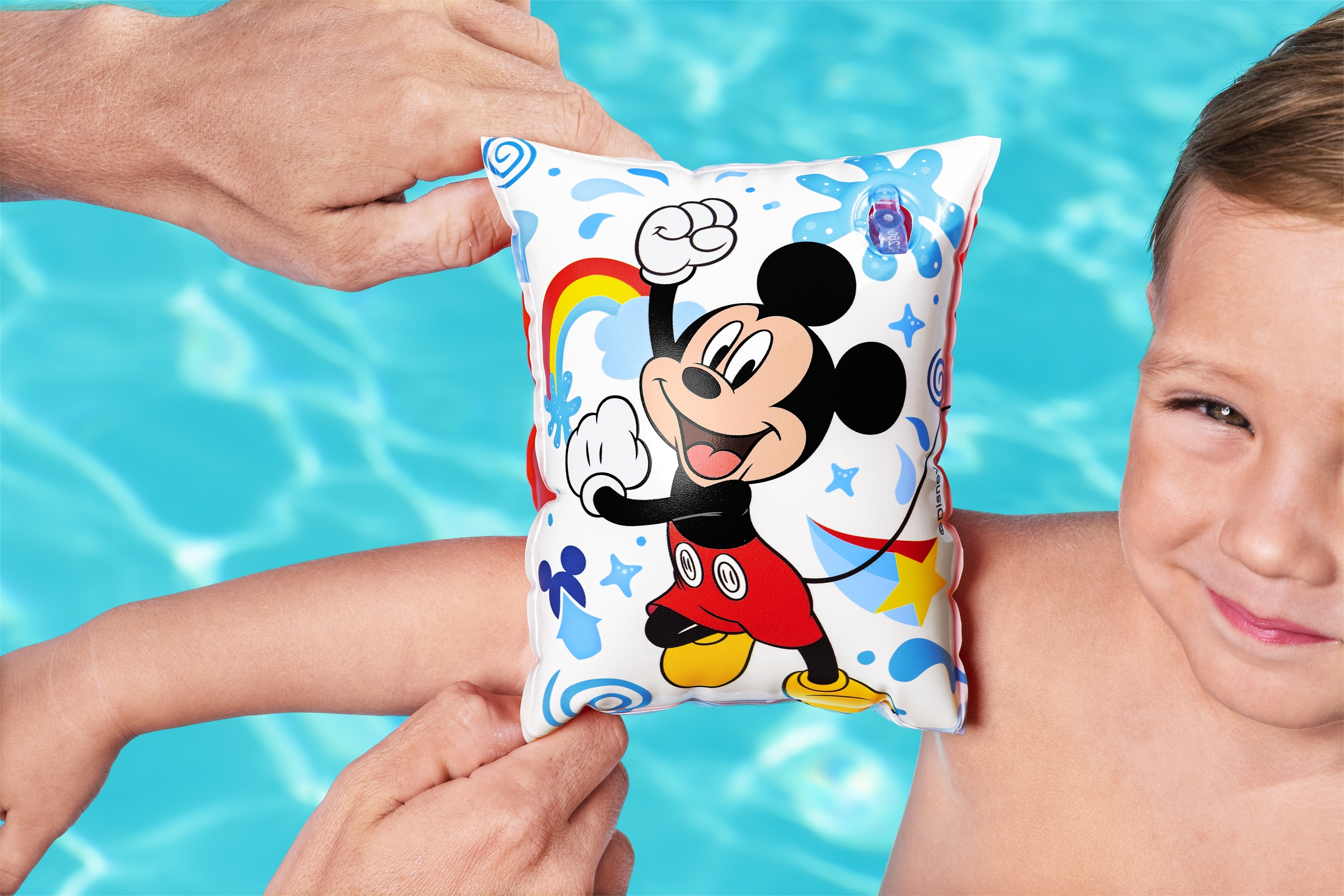 Ein Kind mit einer Mickey-Mouse-Armmanschette im Pool, das von einem Erwachsenen gehalten wird. Das Kind lächelt.