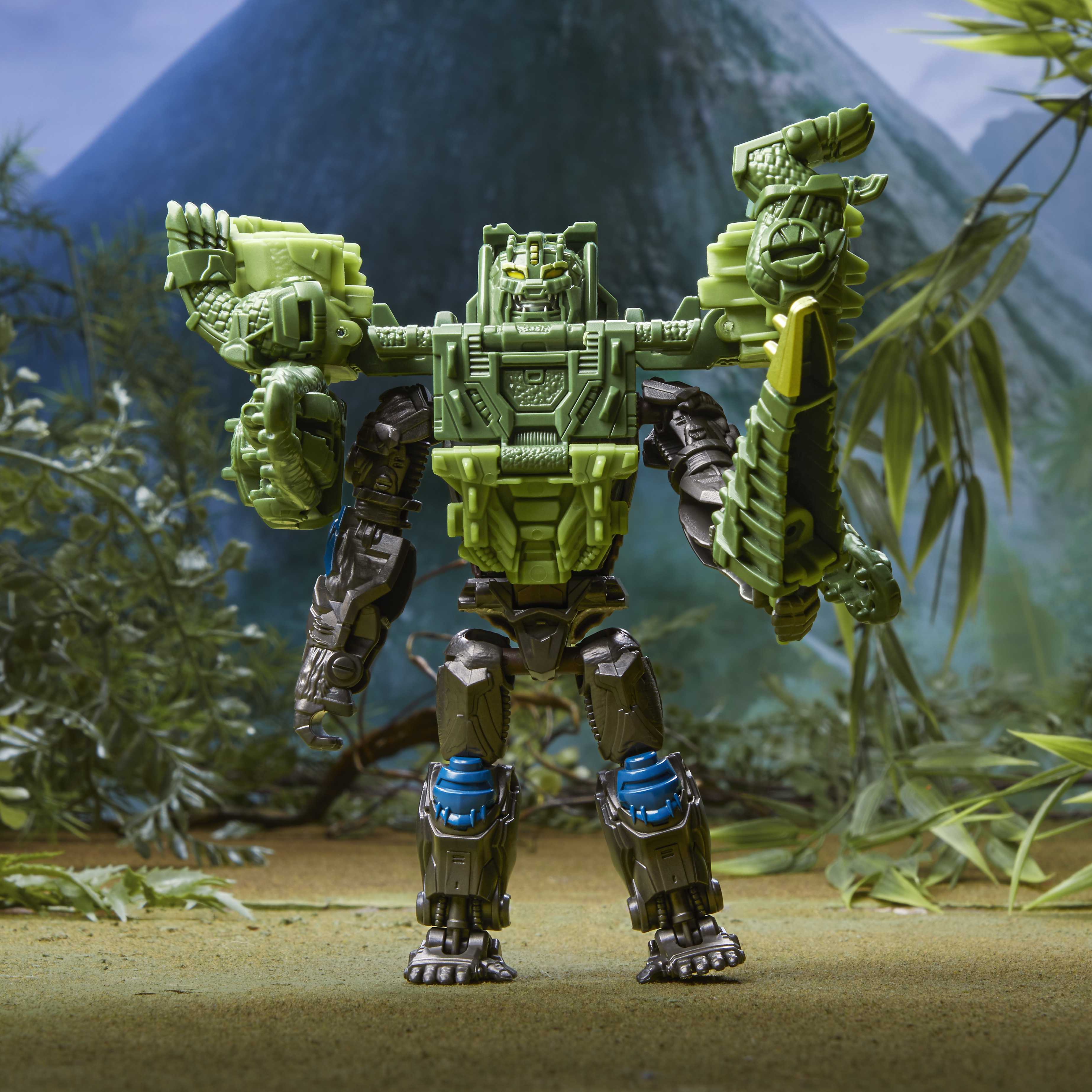 Giocattolo robot verde in piedi. Ha le braccia alzate. Lo sfondo ha una montagna e piante.