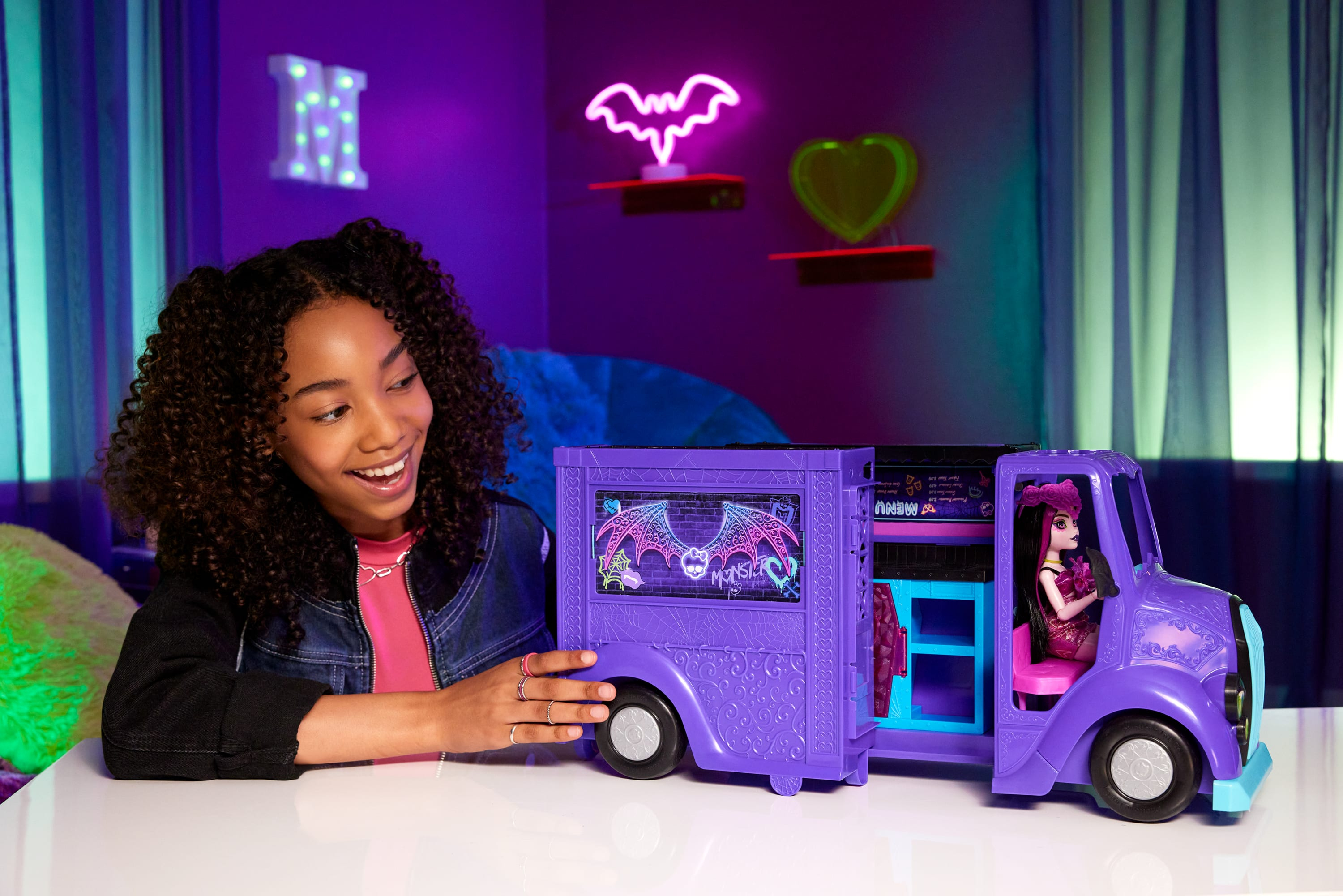 Ein Mädchen lächelt einen lila Spielzeug-Truck mit einer Puppe im Inneren an. Neon-Schilder sind an der Wand.
