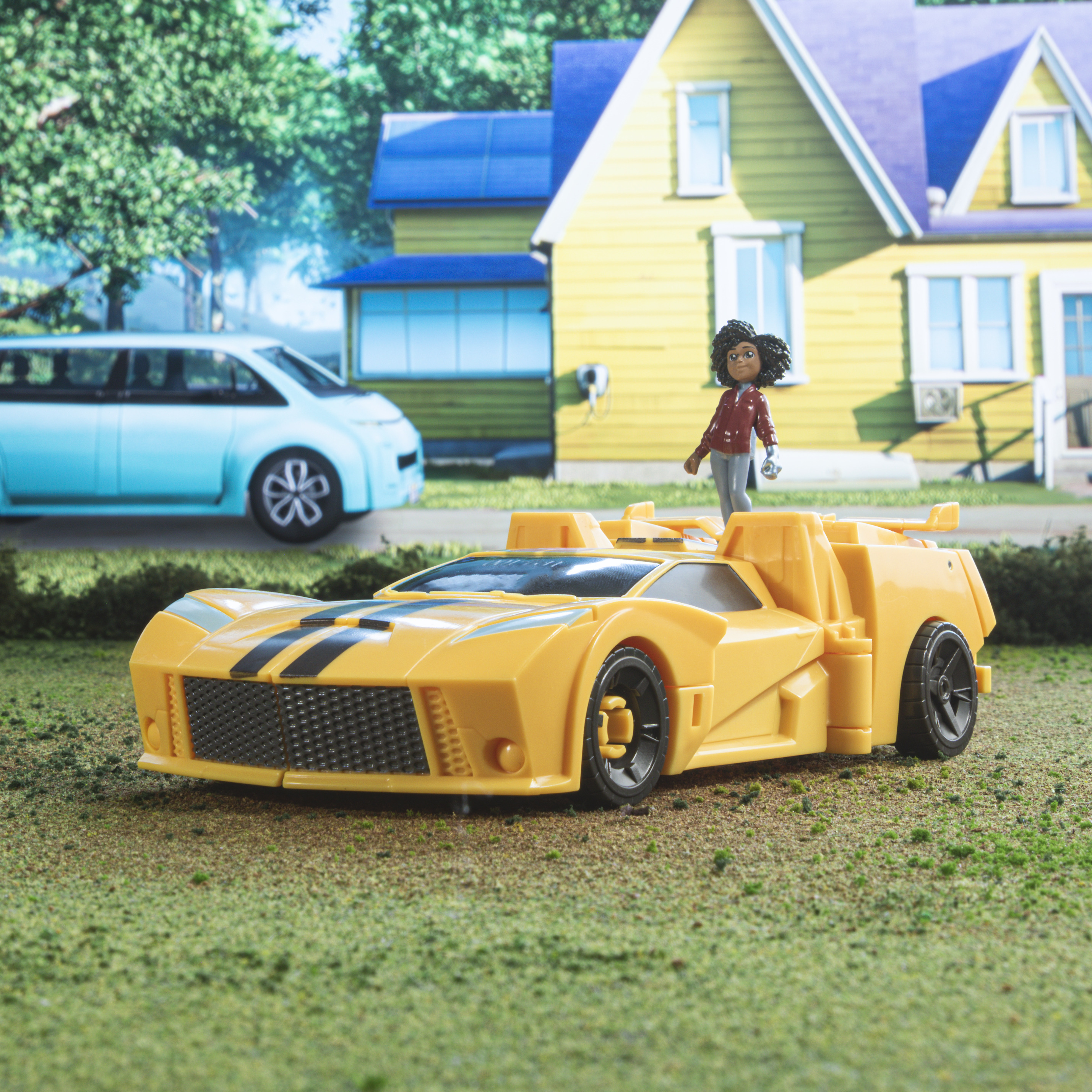 Un coche de juguete amarillo con una figura encima está sobre la hierba. Un coche azul y una casa amarilla están al fondo.