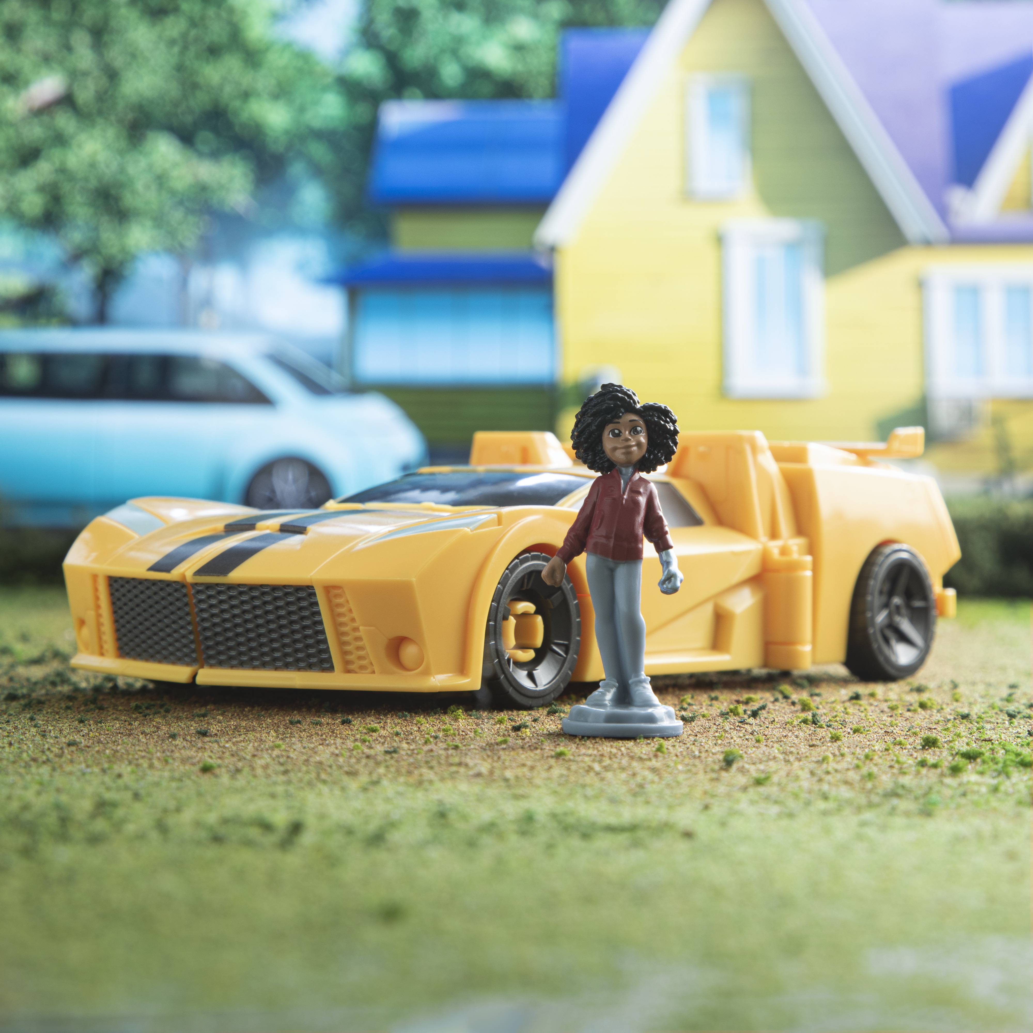 Un coche de juguete amarillo con una raya negra, una figura de juguete y una casa al fondo.