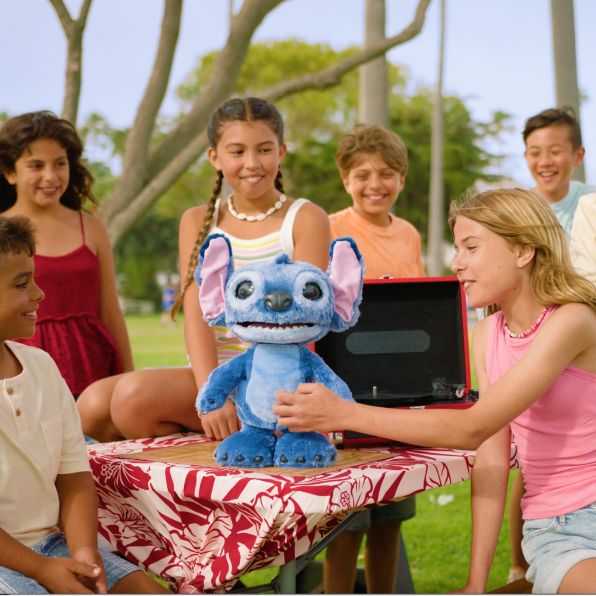 Grupa dzieci otacza pluszową zabawkę Stitch na stole, uśmiechając się. Czerwony gramofon stoi obok zabawki.