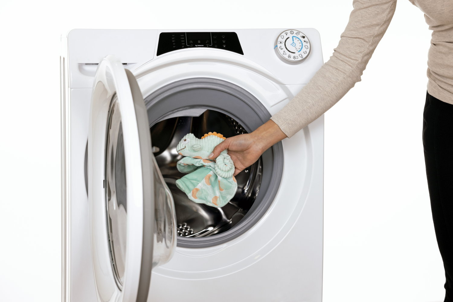 Osoba wkłada zieloną i pomarańczową zabawkę do pralki. Maszyna jest biała, a tło również białe.