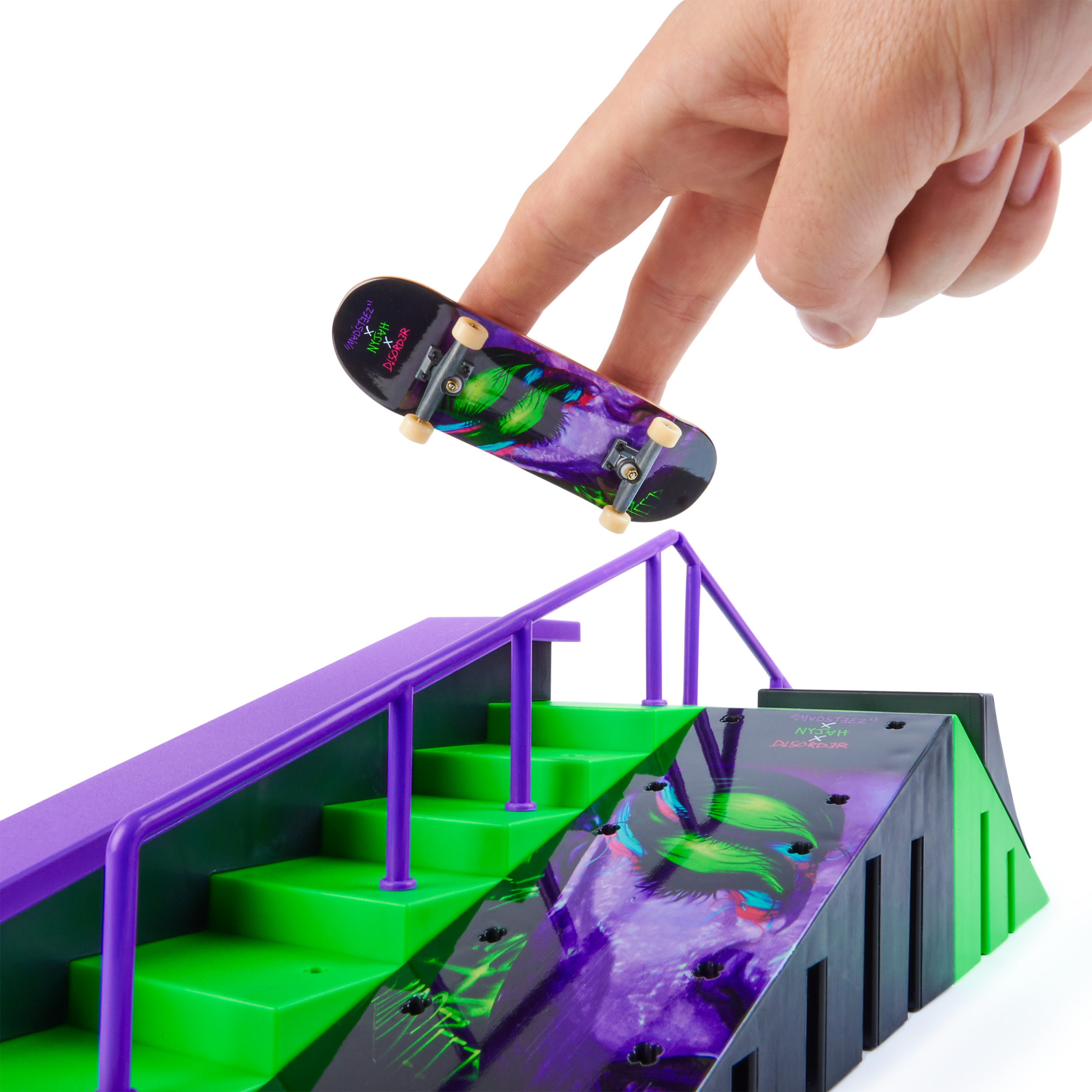 Una mano sostiene una patineta en miniatura sobre una rampa con colores morados y verdes. La tabla tiene un diseño gráfico.