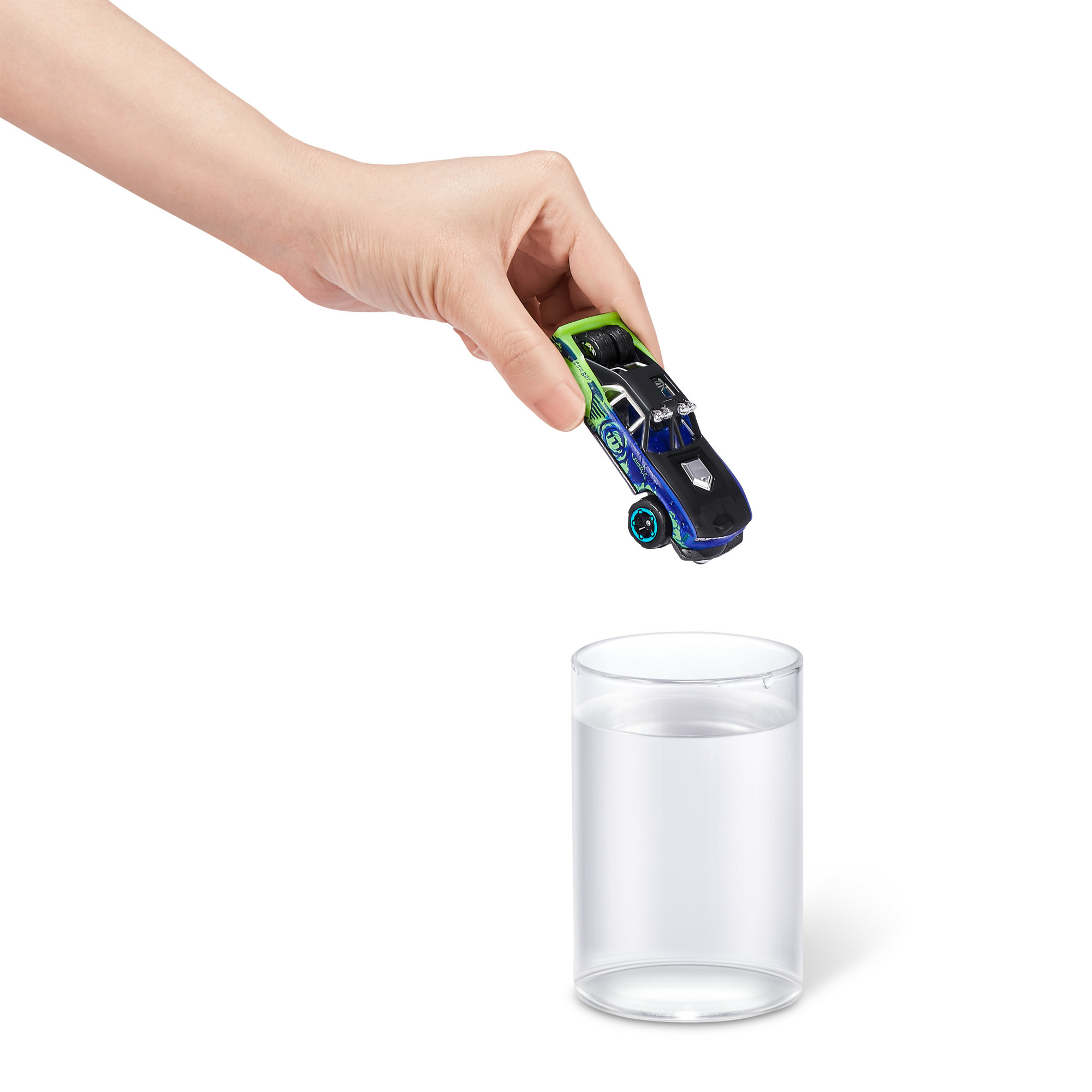 Ręka trzyma zabawkowe auto nad szklanką wody. Samochód jest zielony, czarny i niebieski.