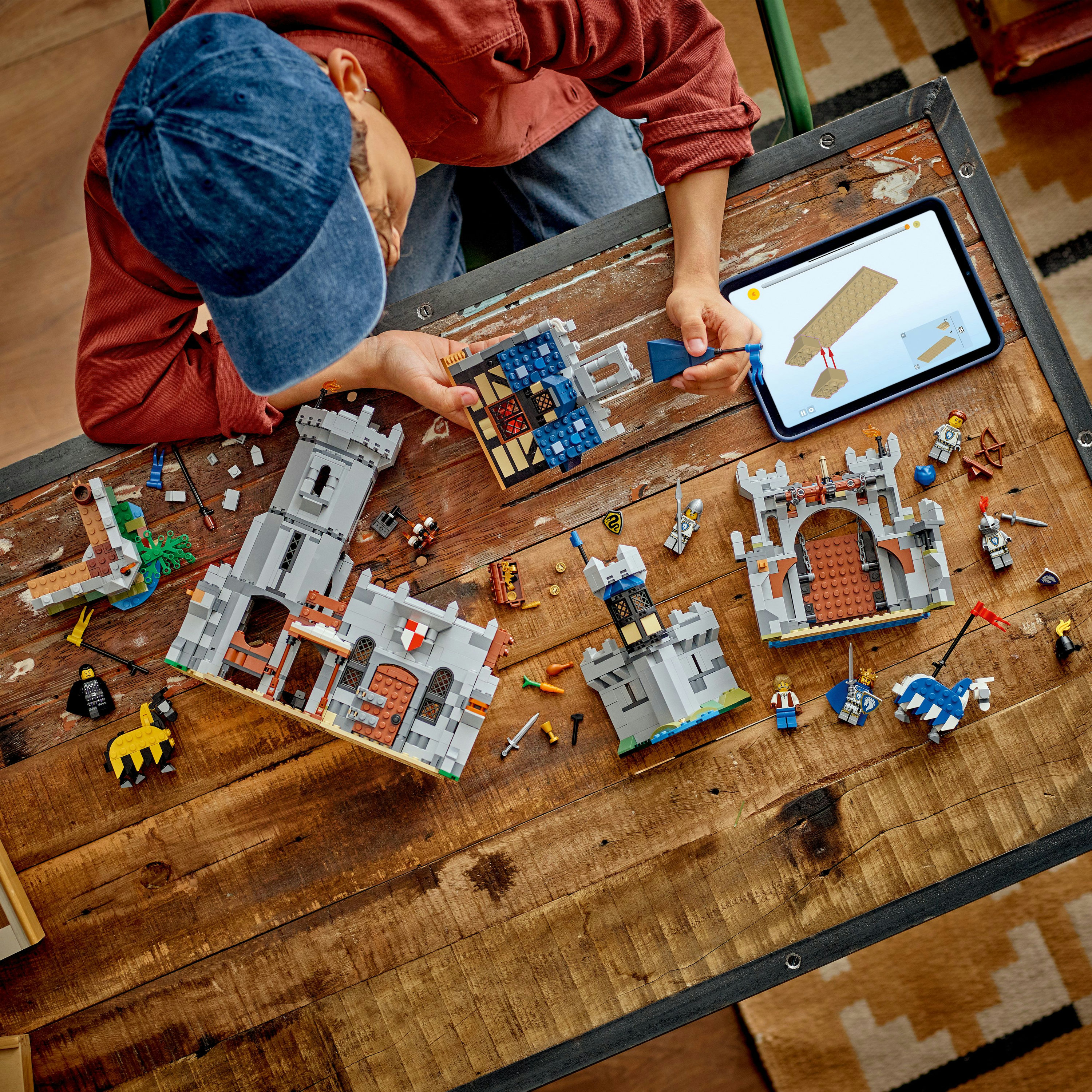 Osoba buduje zamek z Lego. Elementy, tablet i części zamku są rozłożone na drewnianym stole. Widoczna jest niebieska czapka.