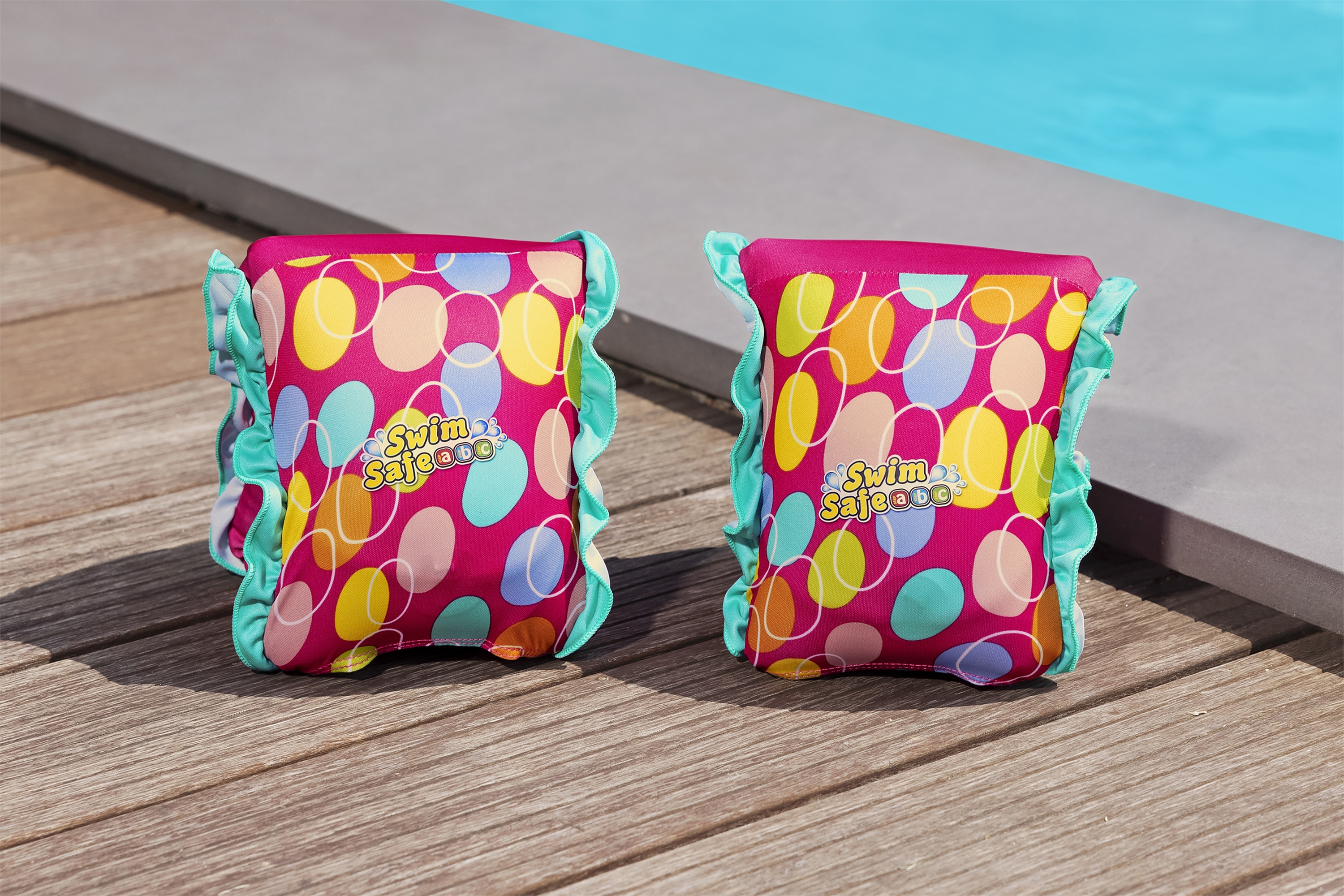 Zwei rosa Schwimmflügel mit bunten Punkten, Text: Swim Safe. Sie liegen auf Holzplanken, im Hintergrund ein Pool.