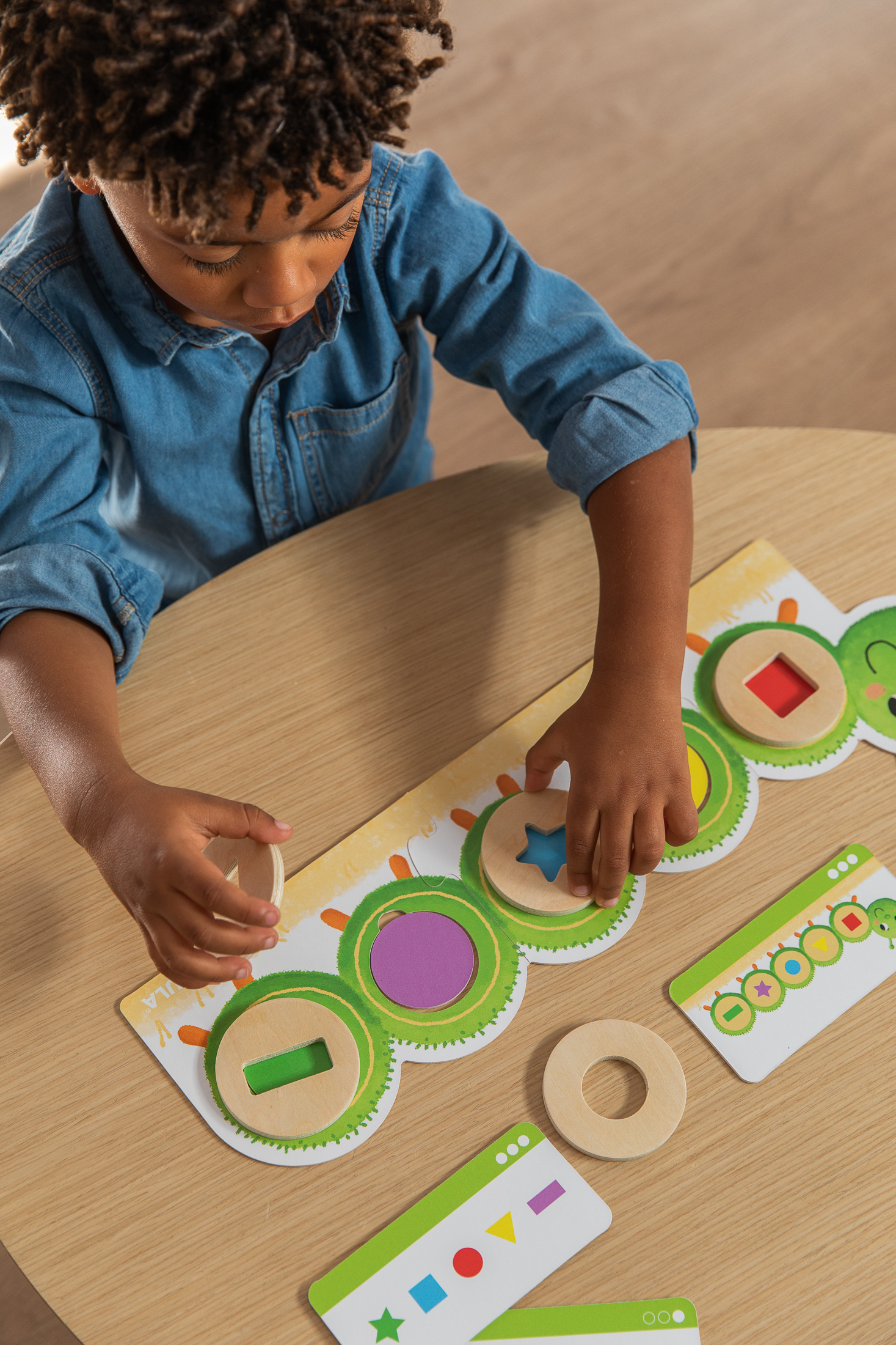 Un niño con camisa de mezclilla juega con un juguete de madera para emparejar formas sobre una mesa de madera. El juego tiene formas coloreadas.