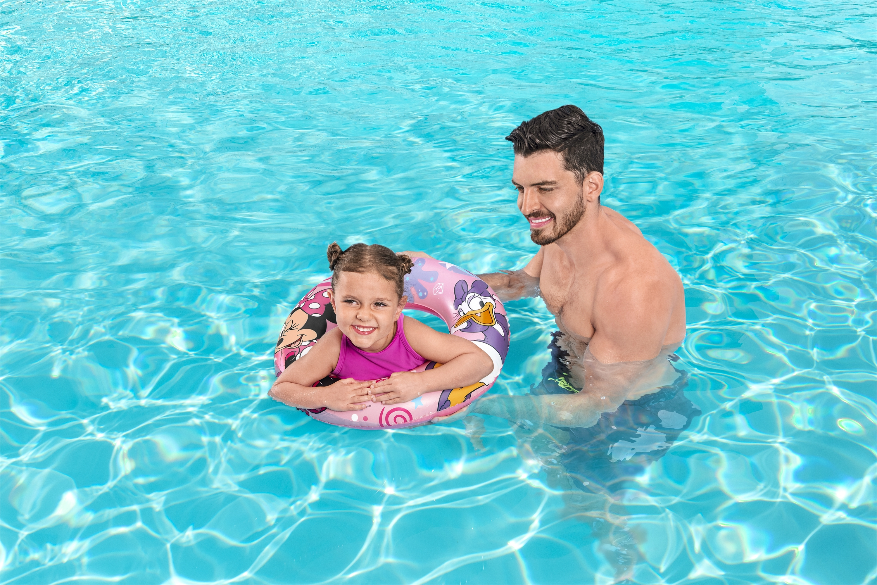 Una niña en un flotador rosa sonríe en una piscina con un hombre. El agua es azul. El flotador tiene personajes de dibujos animados.