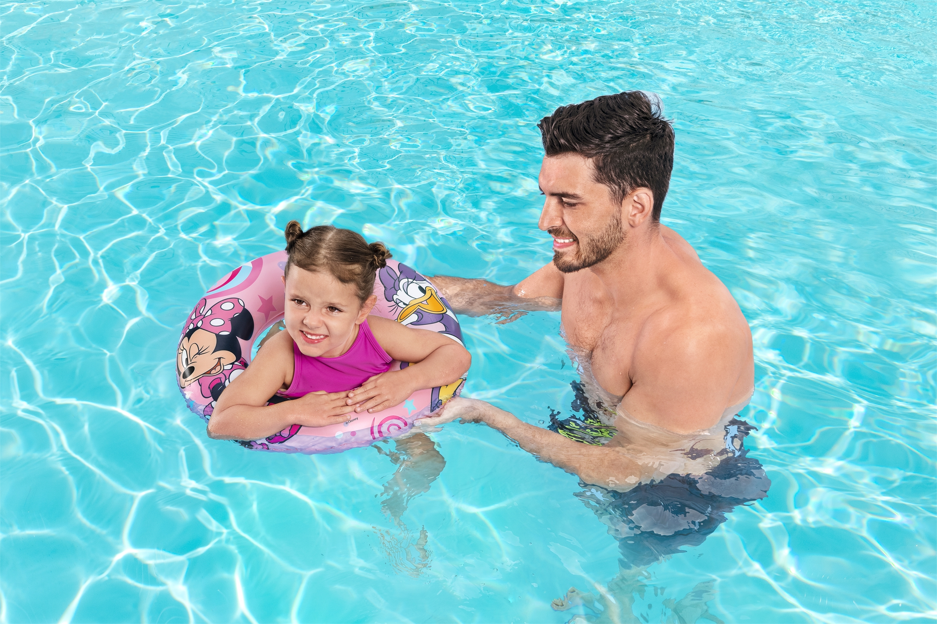 Un hombre ayuda a una niña en un flotador de Minnie Mouse en una piscina. El agua es azul claro.