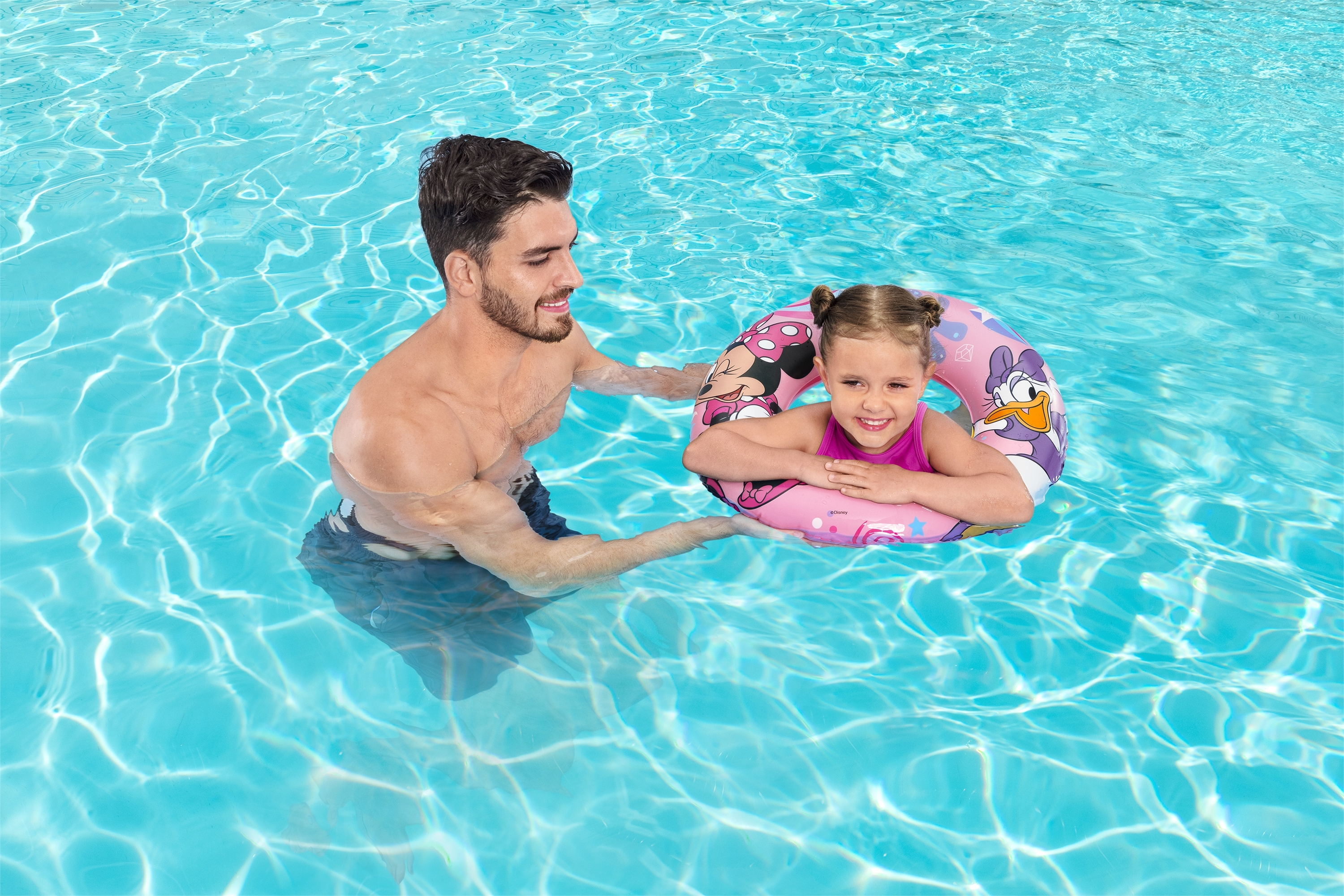 Un hombre ayuda a una niña en un flotador rosa en una piscina. El flotador tiene a Minnie Mouse y Daisy Duck.