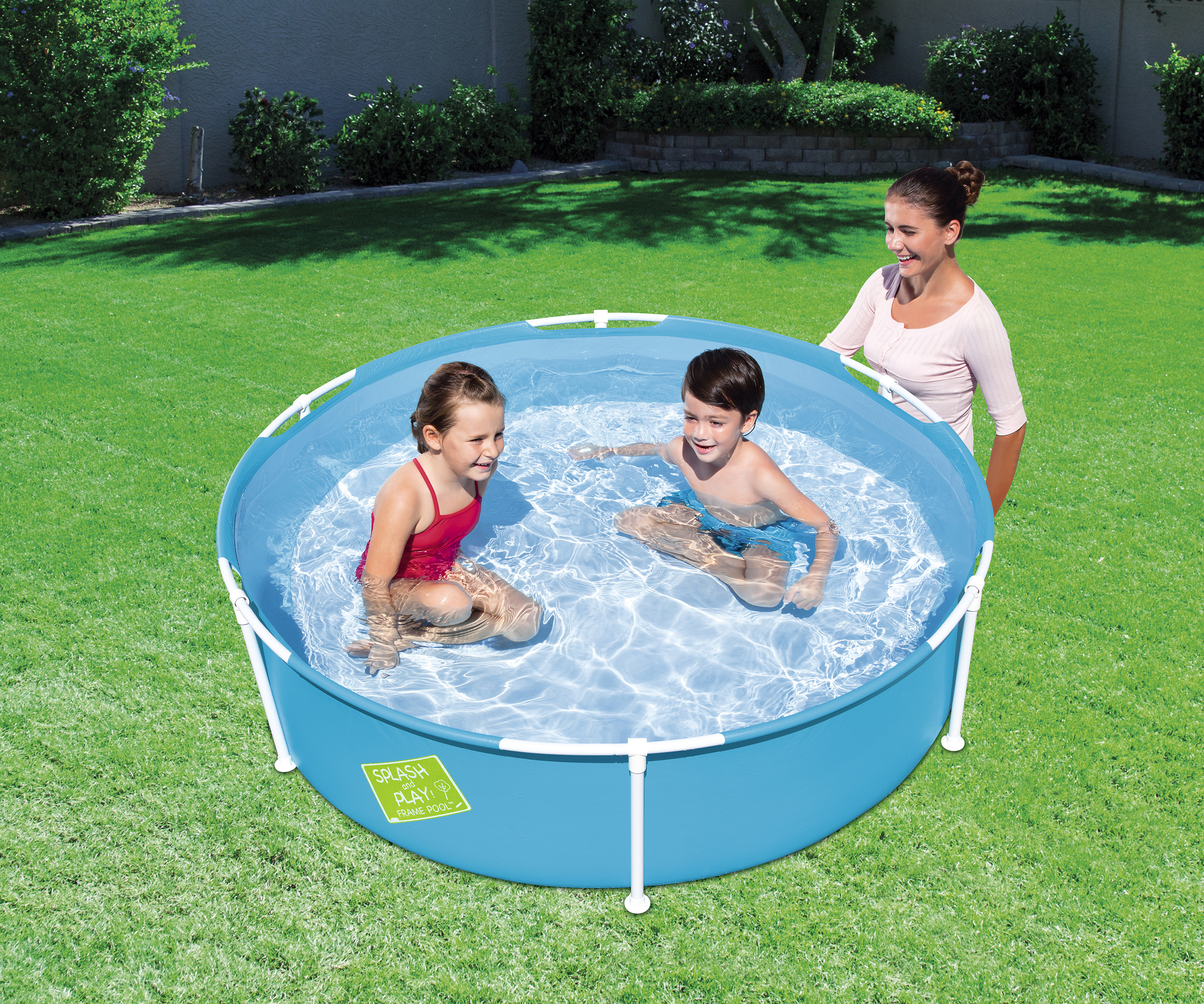 Zwei Kinder in einem blauen Pool, eine Frau steht daneben. Grünes Gras und Büsche im Hintergrund.