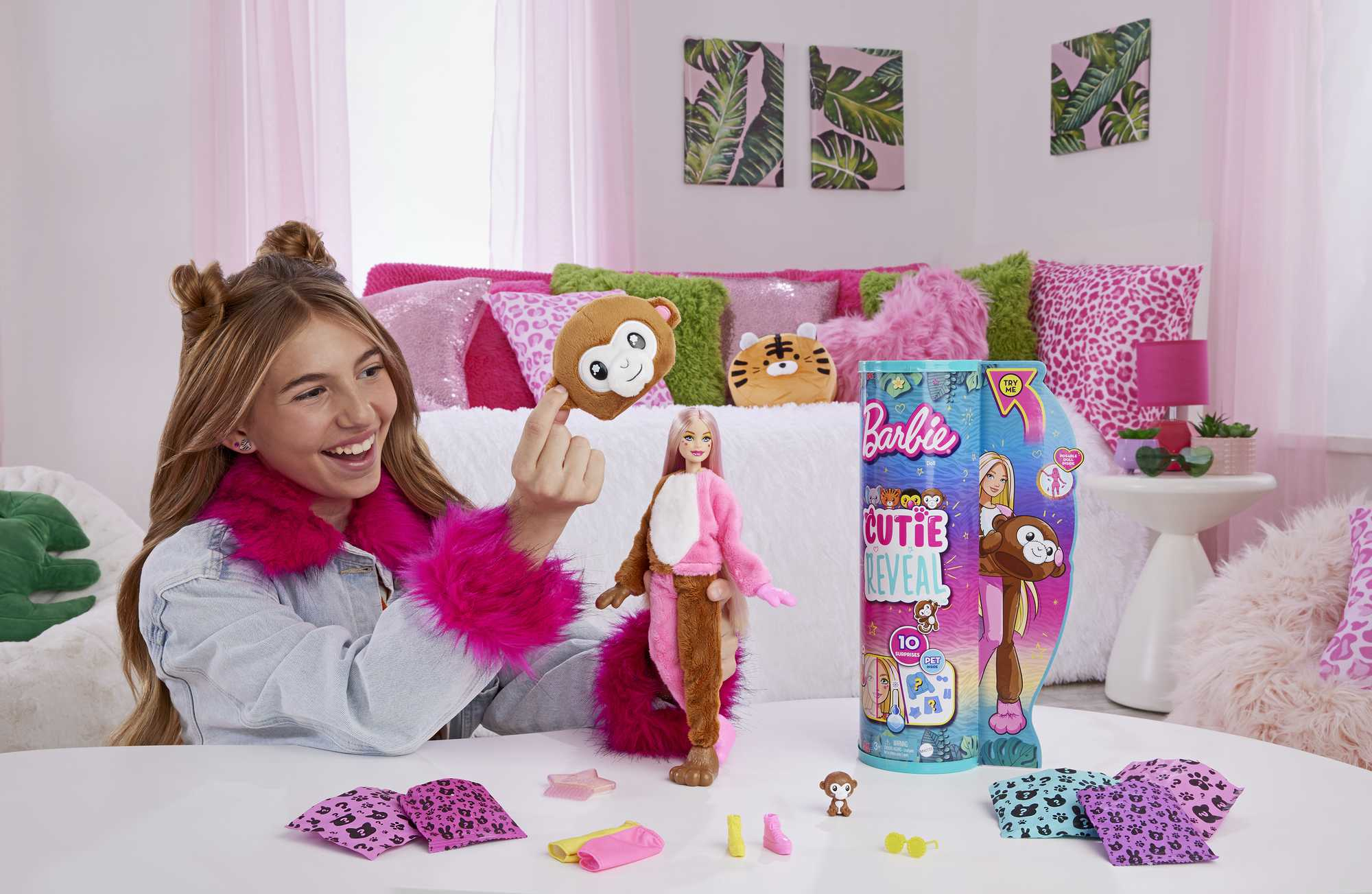 Ein lächelndes Mädchen hält ein Affenplüschtier. Eine Barbie-Puppe, die als Affe verkleidet ist, sitzt auf dem Tisch, mit Zubehör und einer Barbie-Box.
