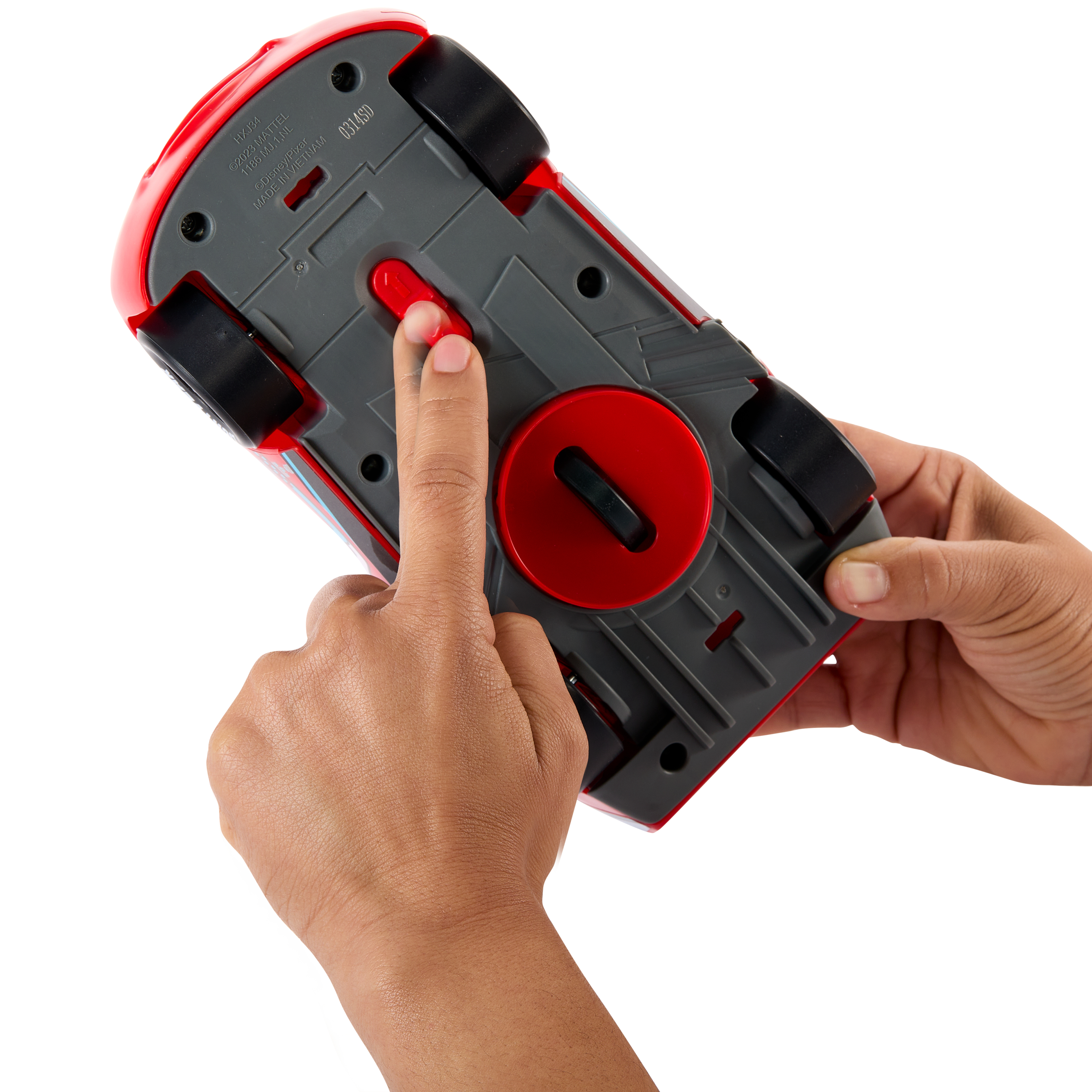 Dłonie osoby naciskają czerwony przycisk na spodzie czerwono-szarego samochodziku. Widoczne są czarne koła.