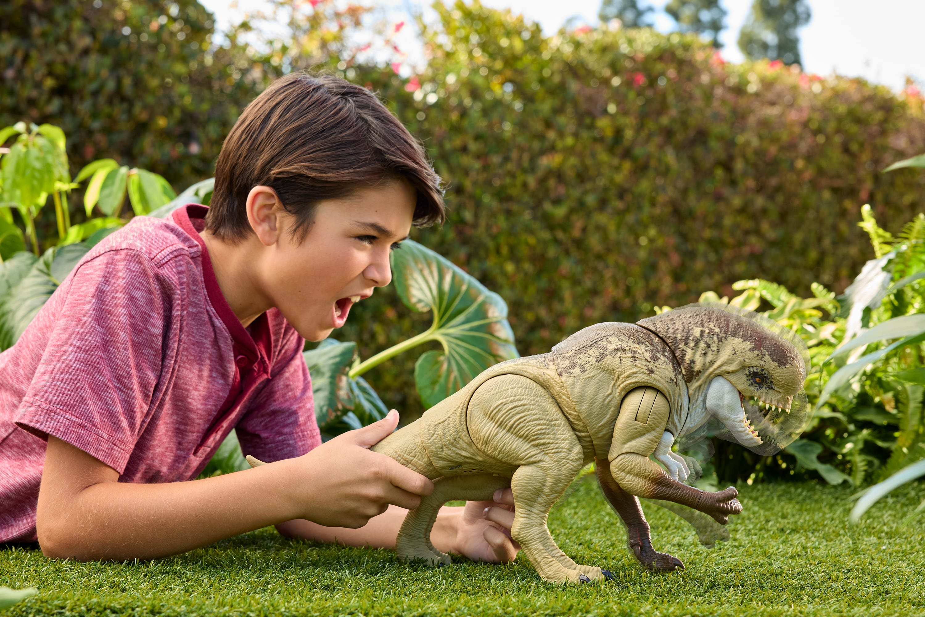 Ein Junge spielt mit einem Dinosaurier-Spielzeug auf Gras. Der Junge ist aufgeregt. Grünes Laub im Hintergrund.