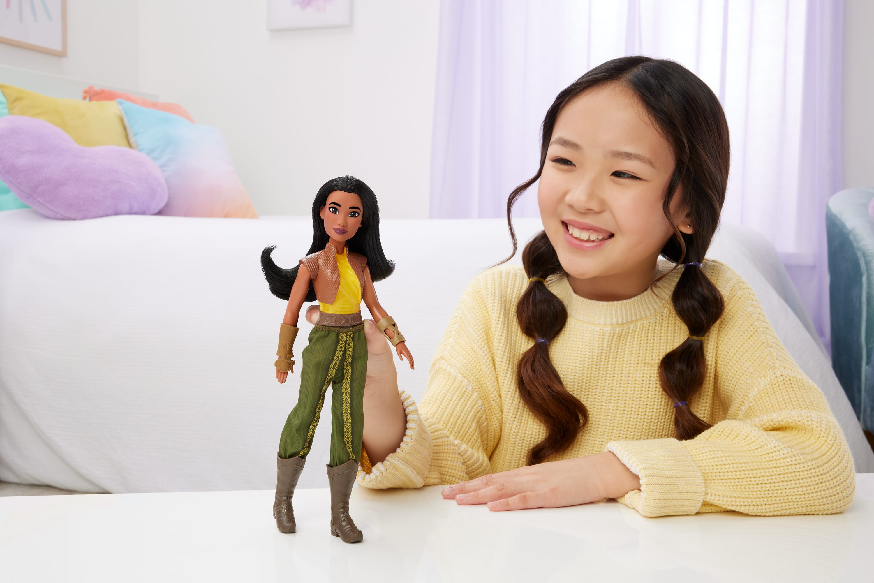 Dziewczyna uśmiecha się do lalki. Lalka stoi na białej powierzchni. W tle widać łóżko i okno.