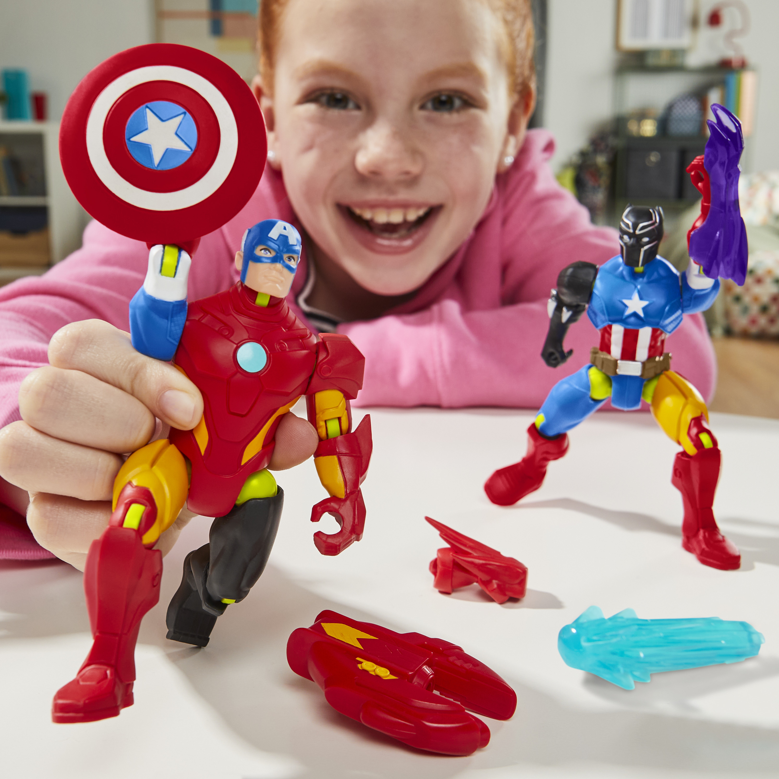 Ein Mädchen lächelt und hält Captain America- und Iron Man-Spielzeuge. Weitere Spielzeuge liegen auf dem weißen Tisch. Der Hintergrund ist unscharf.