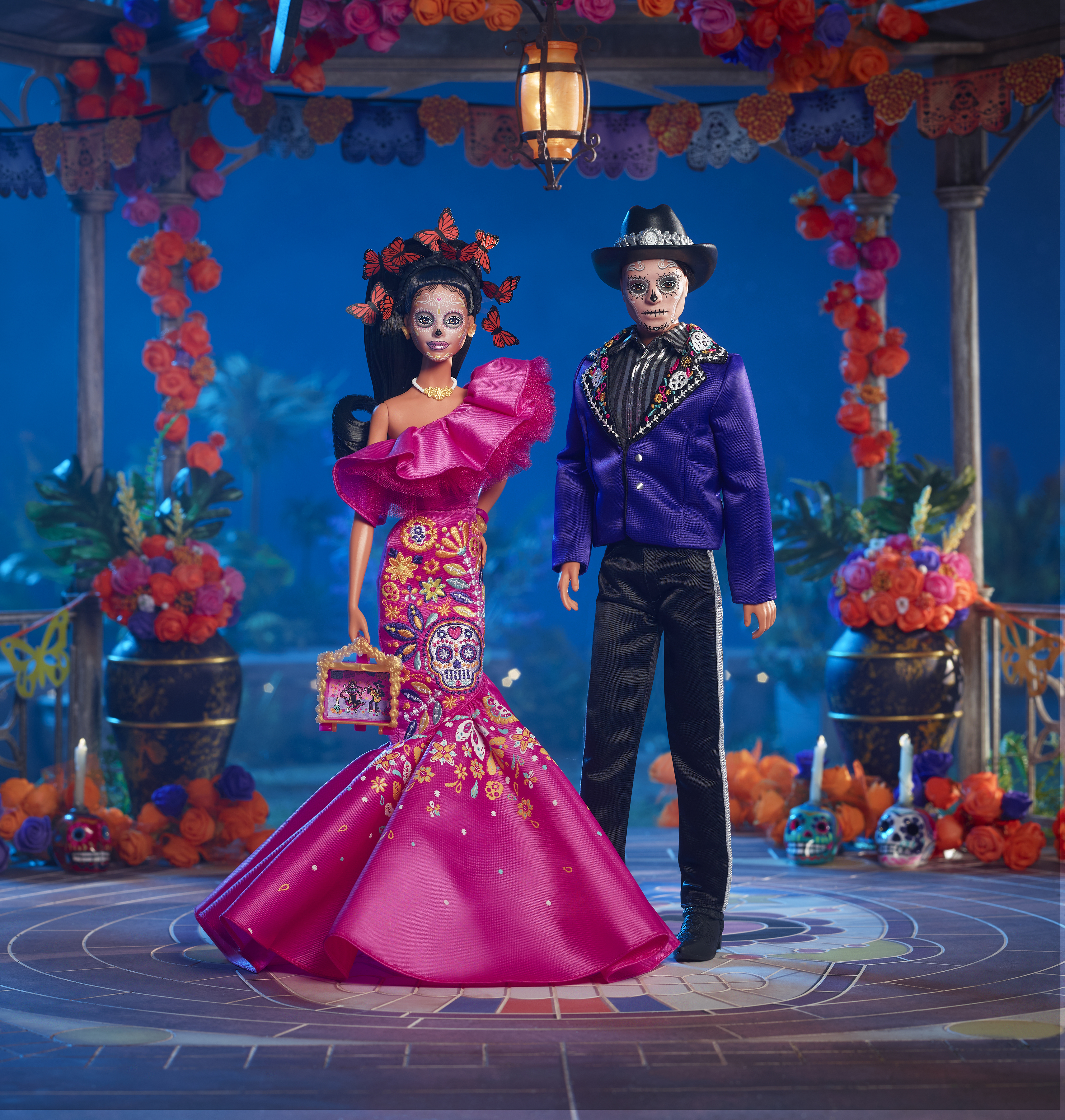 Dwie lalki w strojach Día de Muertos na scenie. Kobieta ma na sobie róż, mężczyzna fiolet. Ozdobne dekoracje.