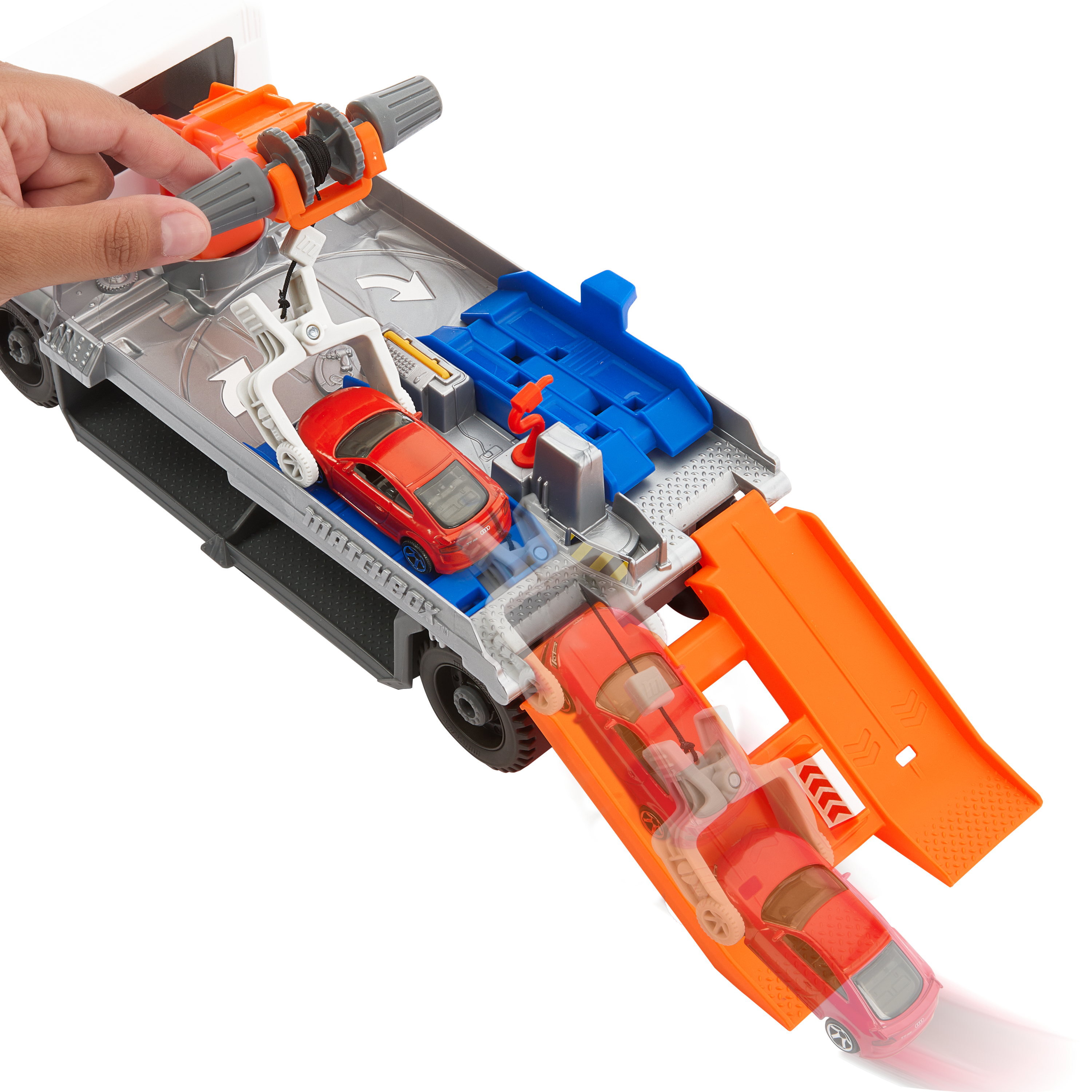 Zabawkowy transporter samochodowy z dwoma czerwonymi samochodami. Ręka obraca szarą i pomarańczową wciągarkę.