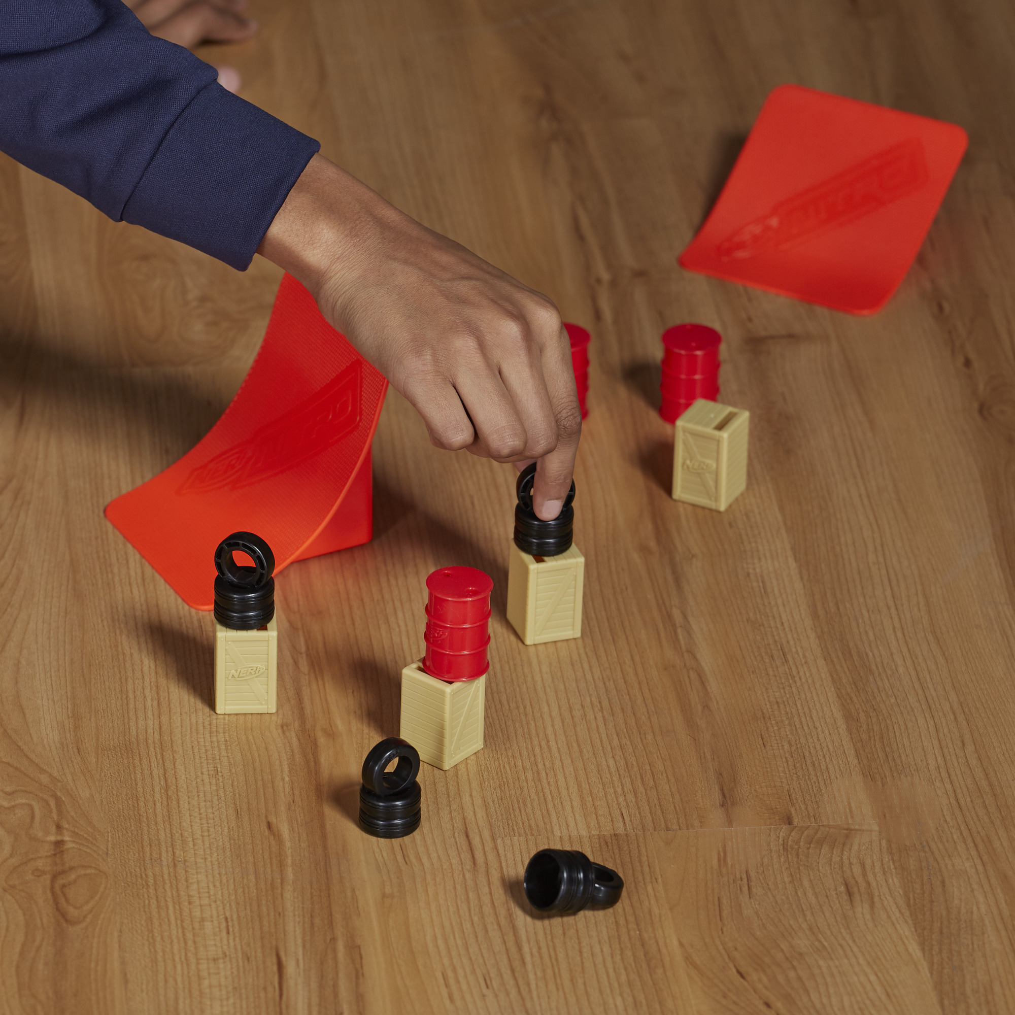 Dłoń wchodzi w interakcję z zabawkami w postaci beczek i skrzyń na drewnianej powierzchni. Czerwona rampa w tle.