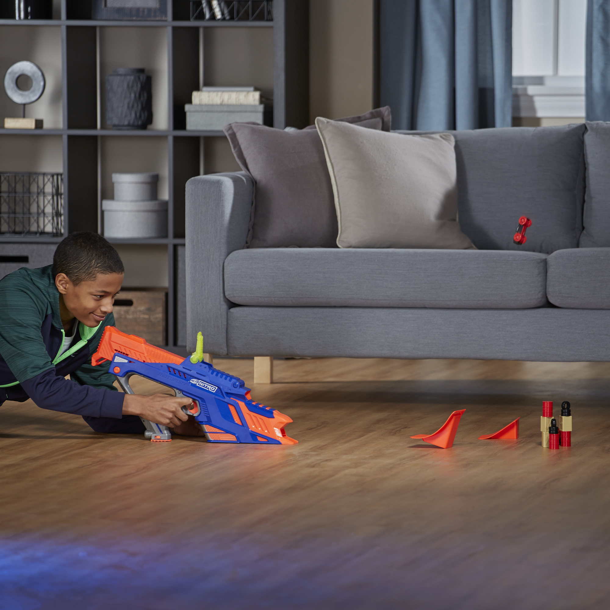 Chłopiec celuje niebiesko-pomarańczową zabawką. Czerwone cele i zabawki samochodowe są na podłodze i kanapie.