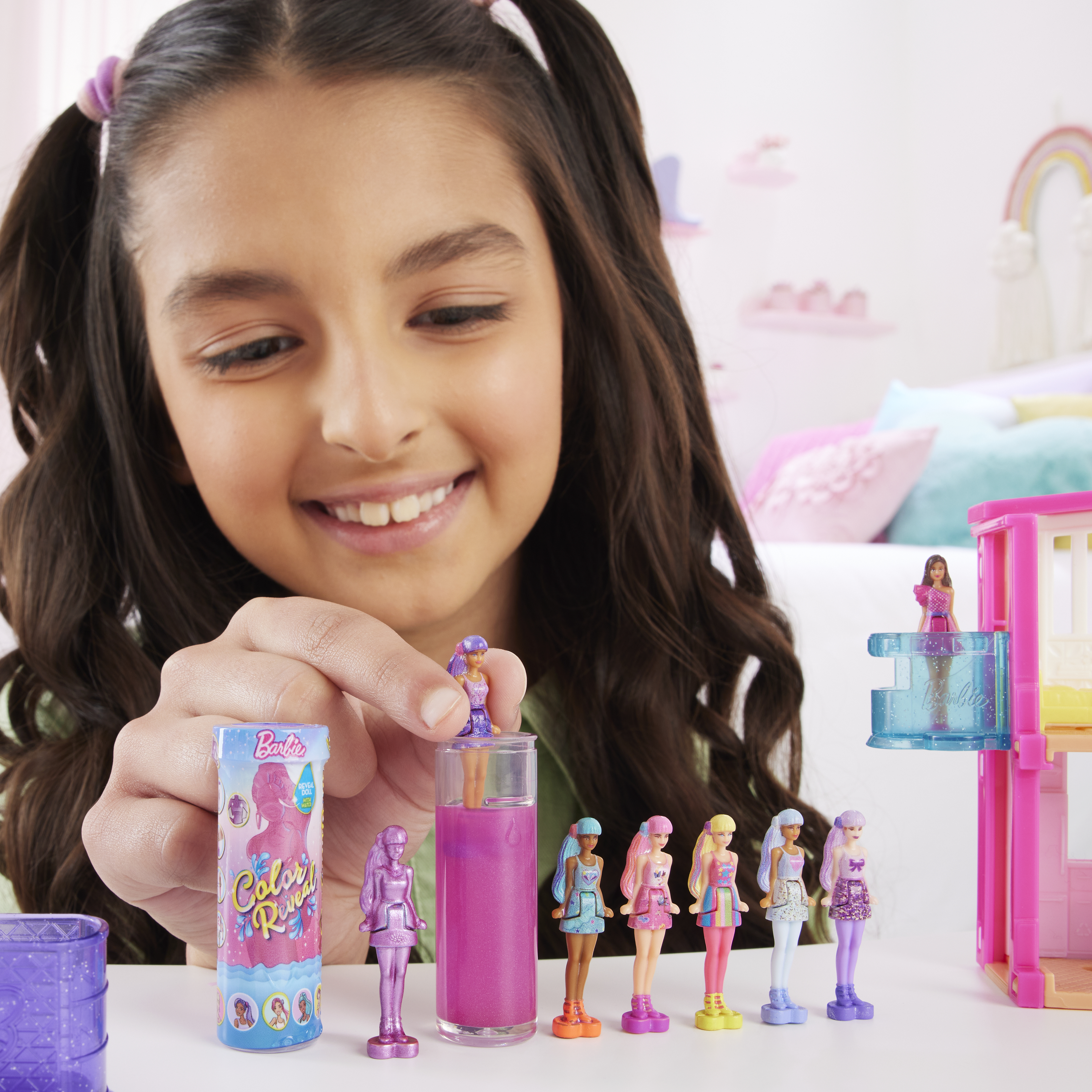 Ein Mädchen lächelt Barbie-Puppen an. Verschiedene Puppen und ein Puppenhaus stehen auf einem weißen Tisch. Ein Regenbogen schmückt den Hintergrund.