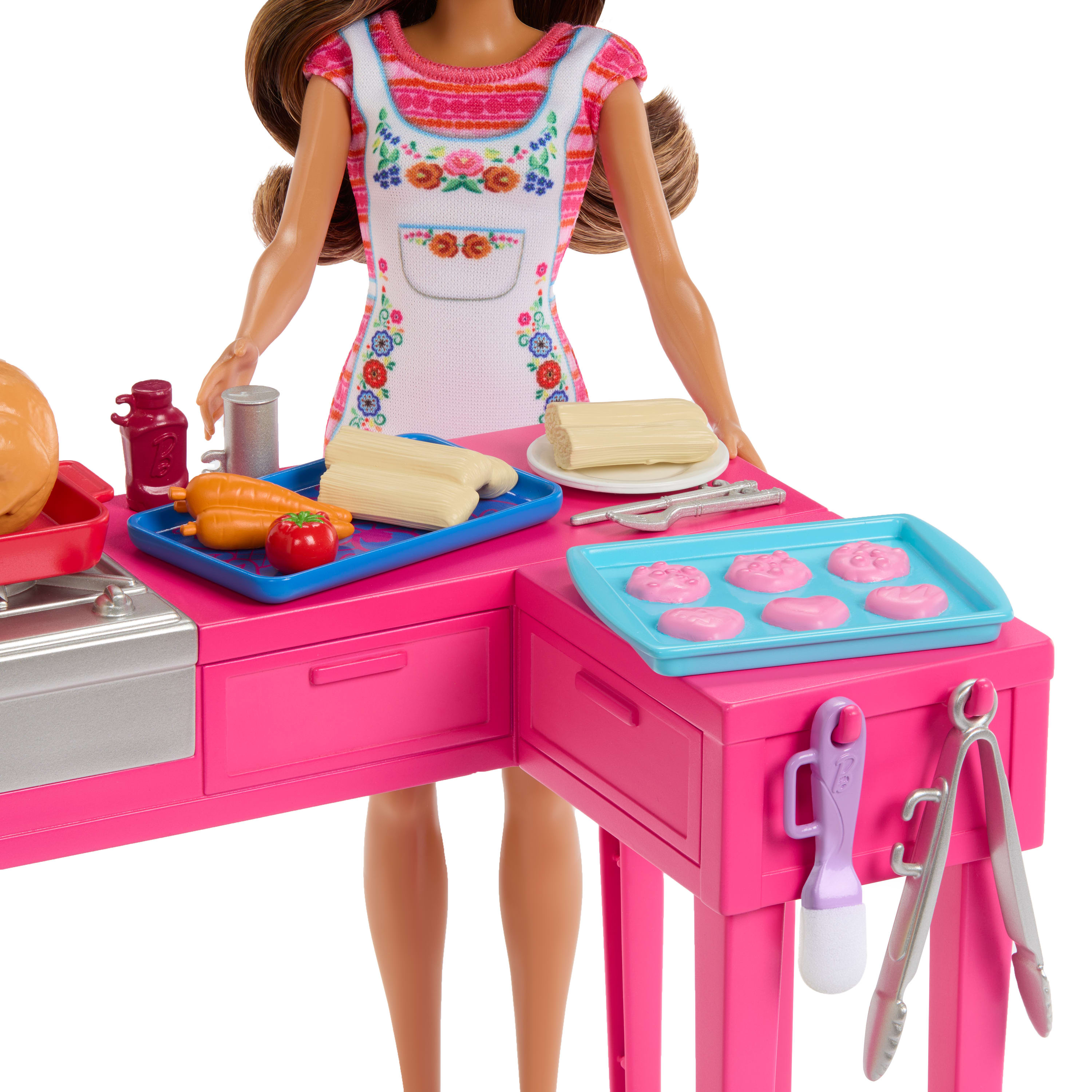 Eine Barbie-Puppe mit Schürze steht hinter einer rosa Spielzeugküche mit Lebensmitteln wie Tamales und Karotten.