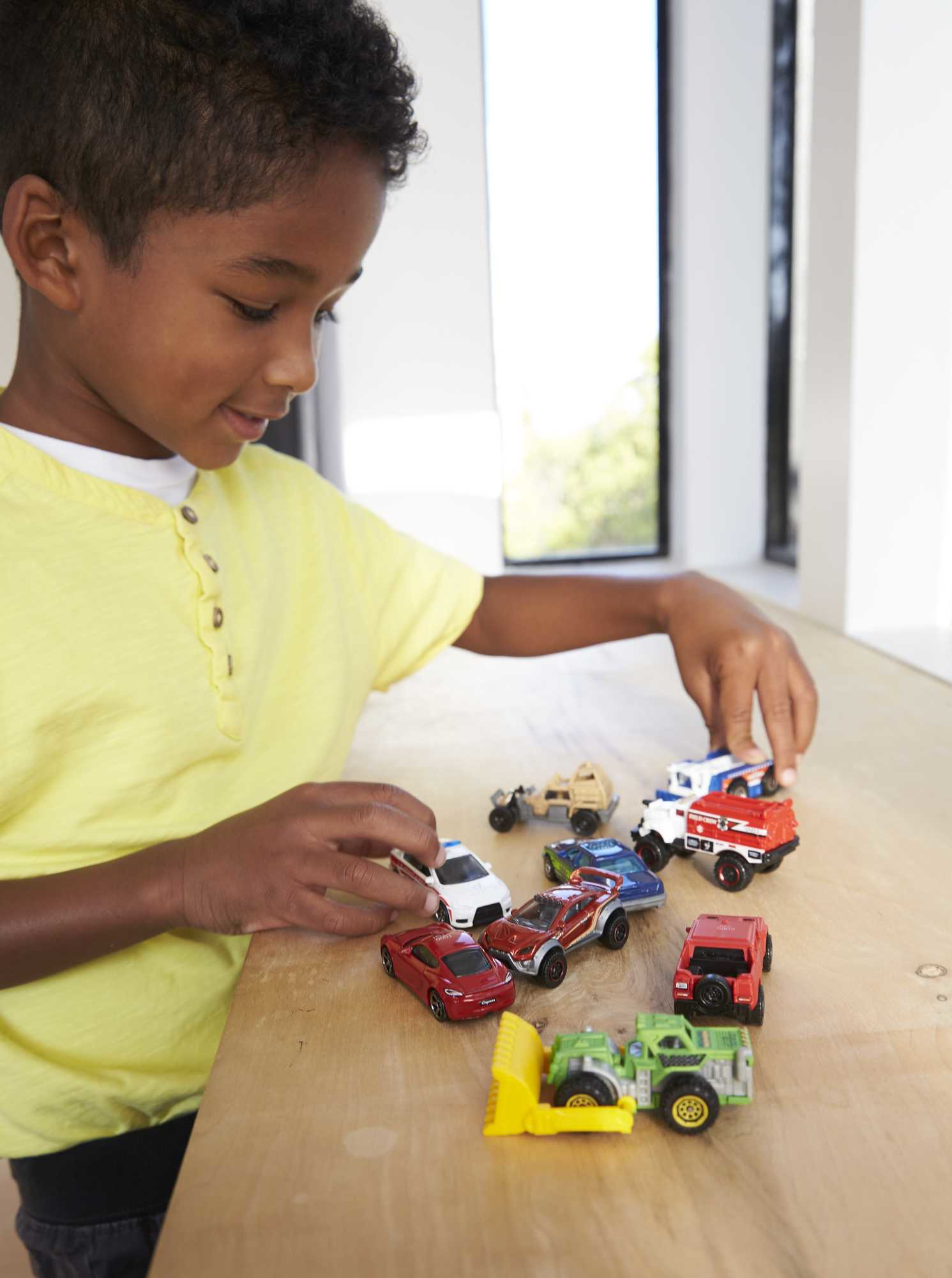 Chłopiec w żółtej koszuli bawi się samochodzikami na drewnianym stole. Samochody są w różnych kolorach i typach.
