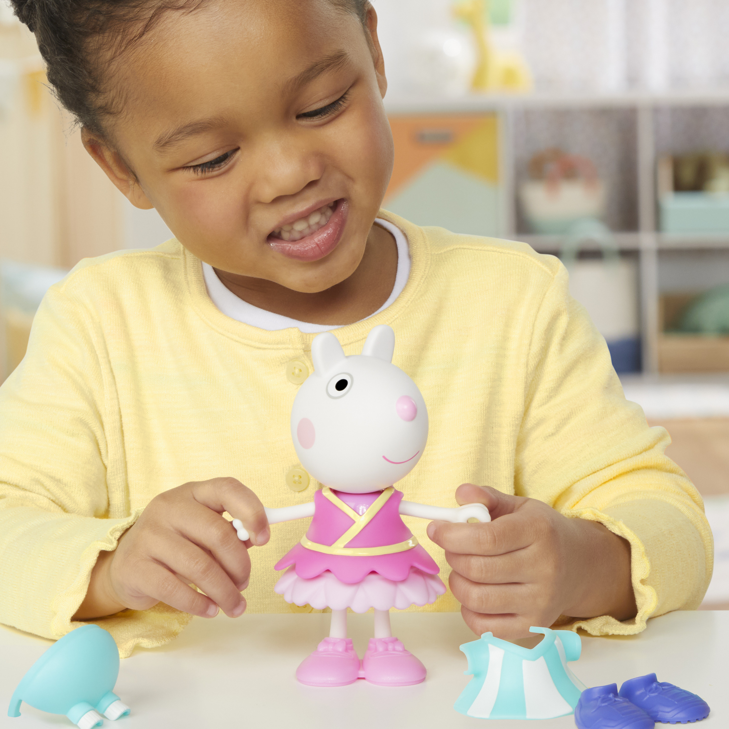 Una niña sonríe a una muñeca con forma de conejito. Hay accesorios como zapatos y ropa en la mesa.