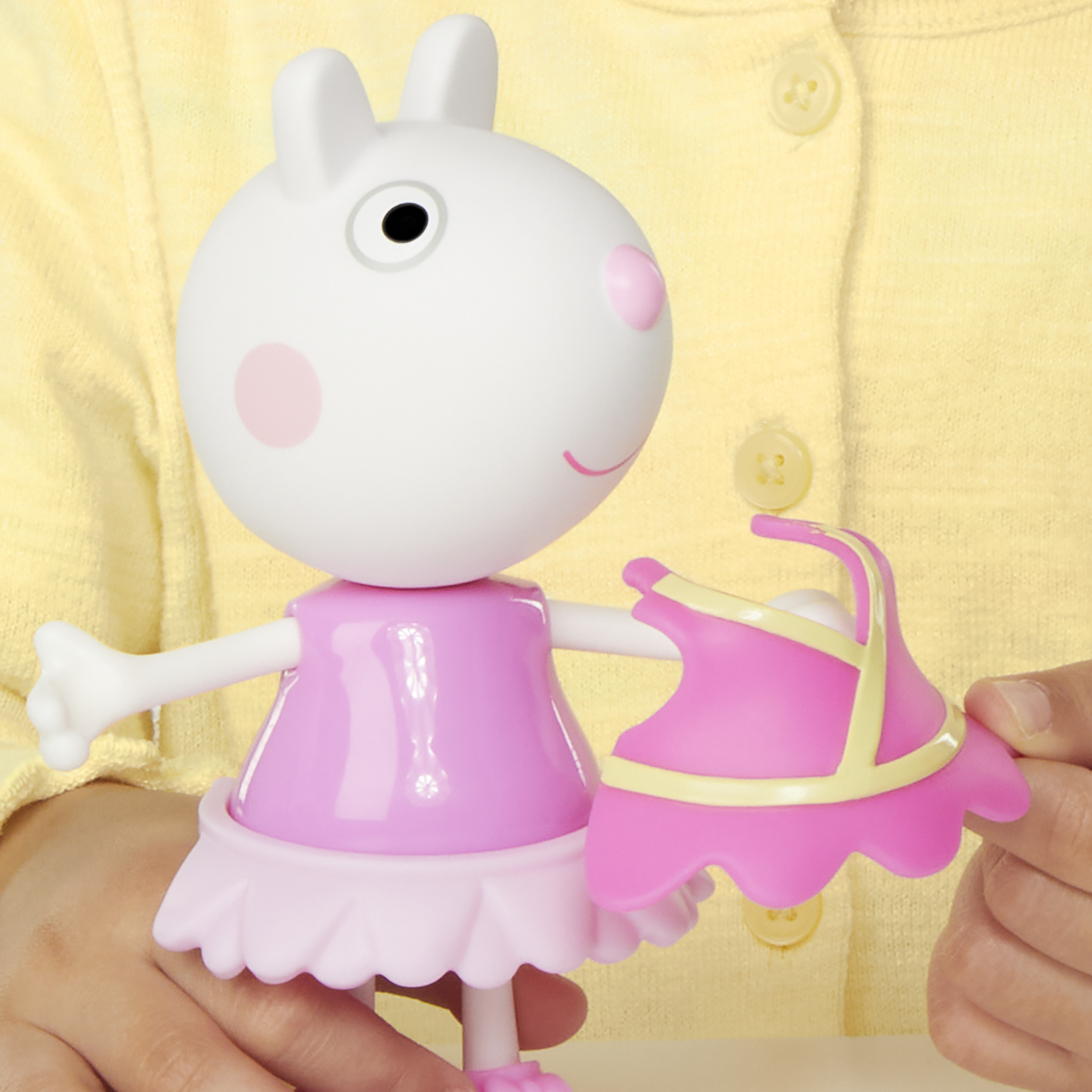 Se sostiene un juguete de Peppa Pig. Es blanco con mejillas rosas y un vestido rosa, sosteniendo un vestido rosa con ribete amarillo.