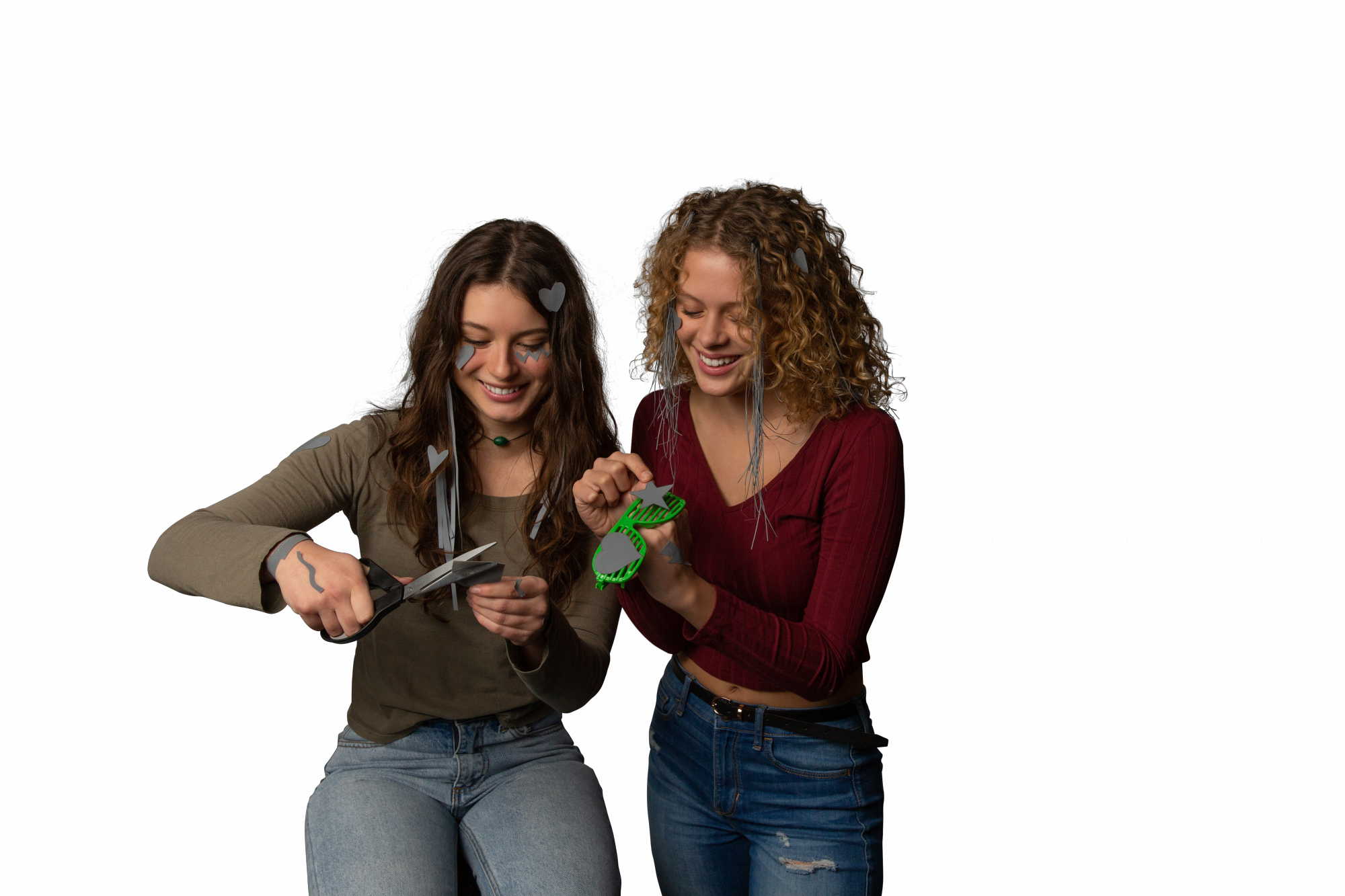 Dwie uśmiechnięte kobiety, jedna tnie papier nożyczkami, druga trzyma zielone okulary w kształcie gwiazdy. Obie mają dekoracje.