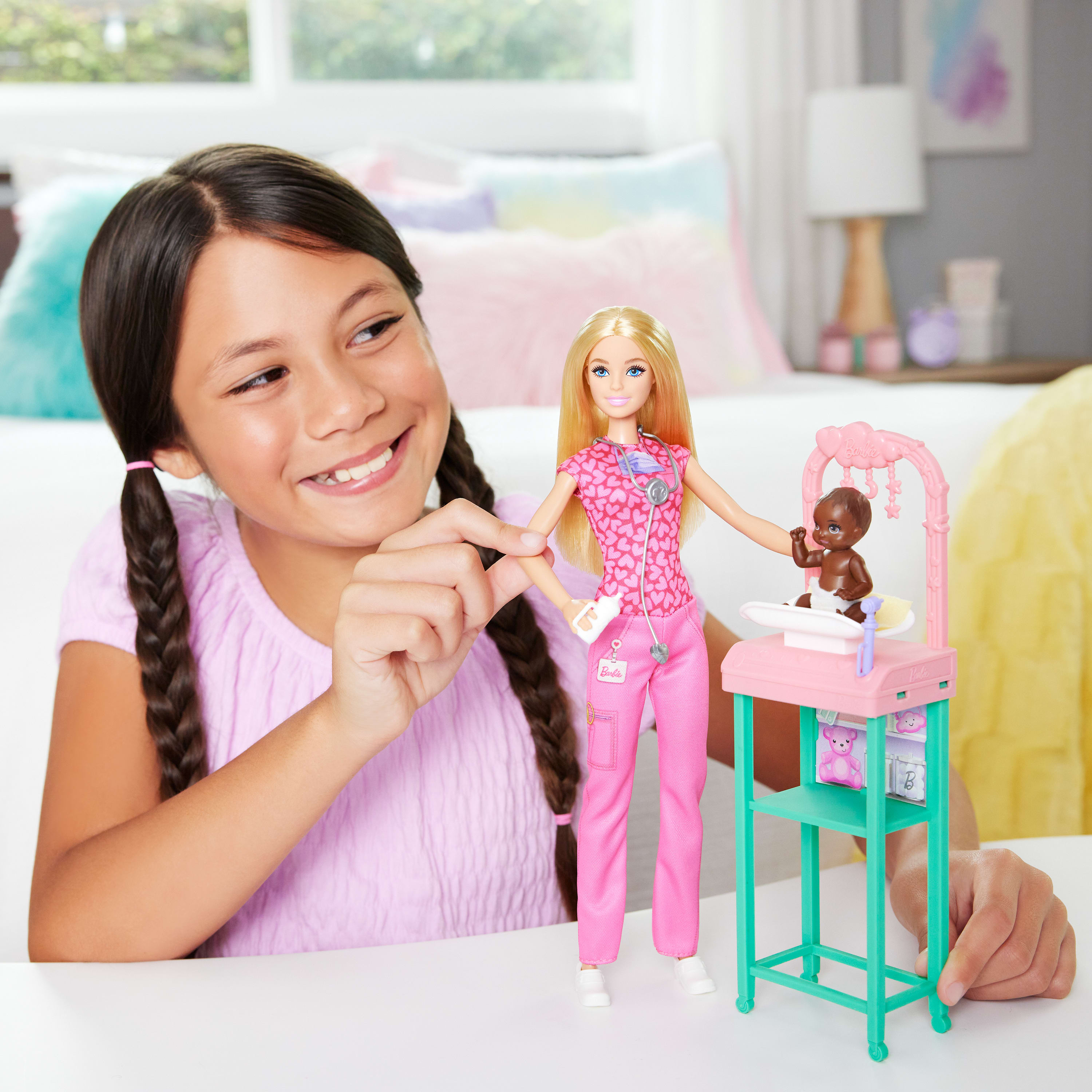 Dziewczyna uśmiecha się do lalki pielęgniarki Barbie i lalki dla dziecka z różową wagą. Tło ma łóżko i lampę.