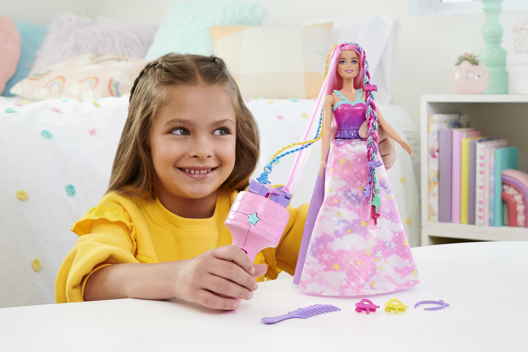 Ein lächelndes Mädchen hält ein rosa Haarstyling-Werkzeug neben einer Barbie-Puppe mit langen, rosa Haaren. Verschiedenes Zubehör auf dem Tisch.
