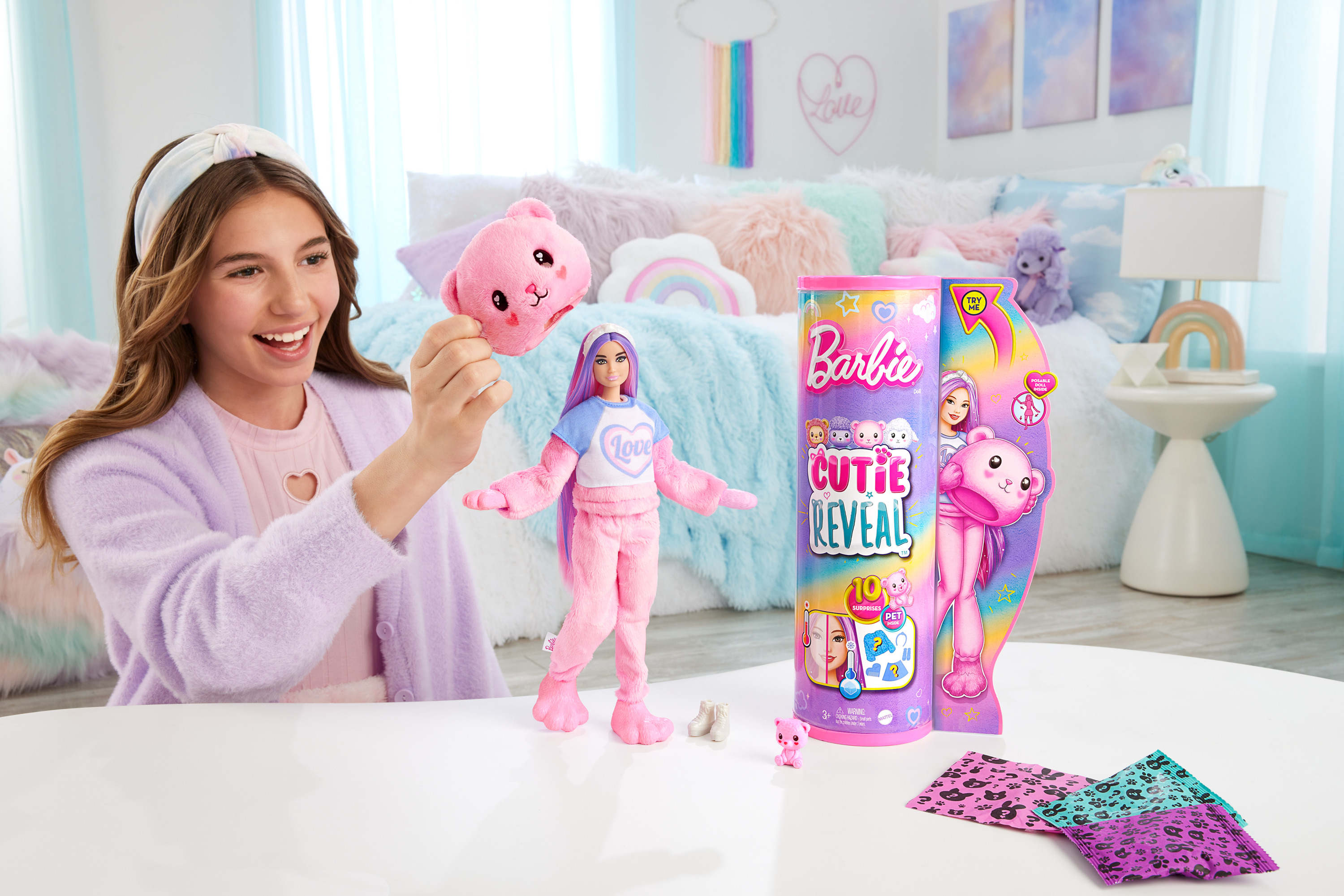 Ein Mädchen hält einen rosa Bärenkopf neben einer Barbie Cutie Reveal-Puppe und -Verpackung auf einem weißen Tisch.